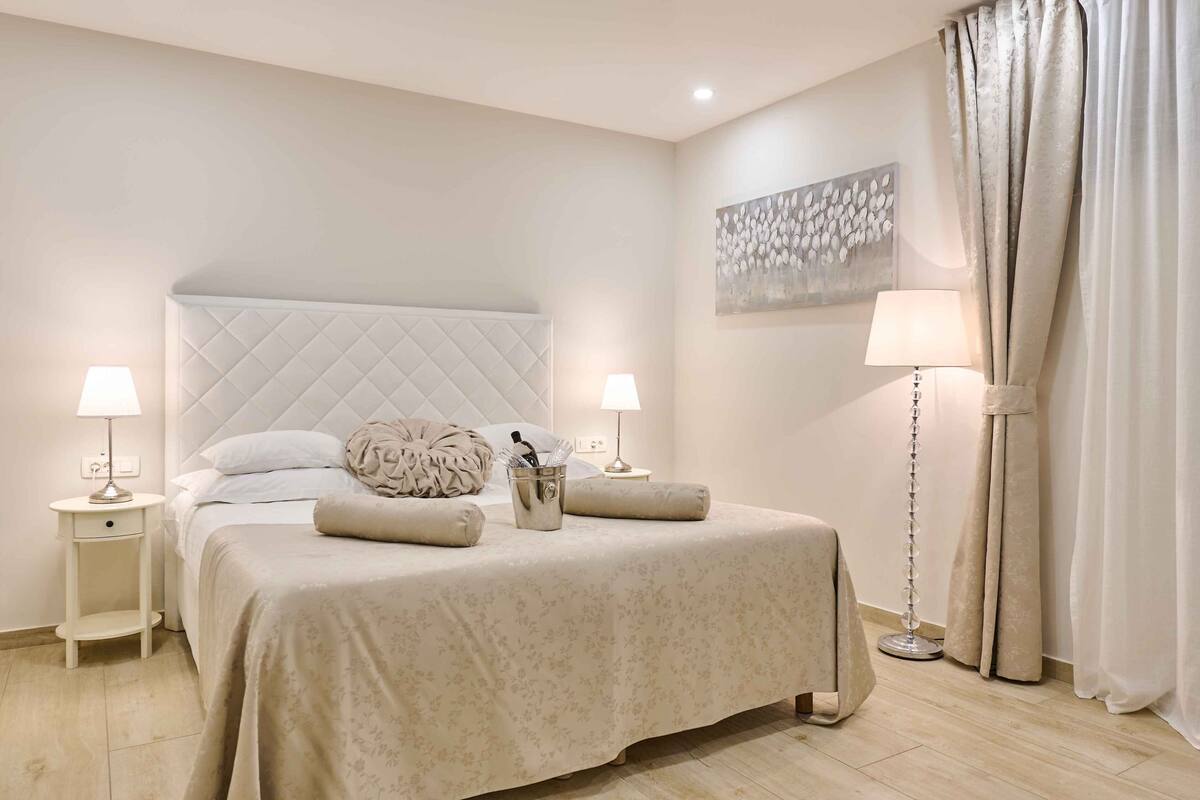 A neatly arranged bedroom with a large bed, two bedside tables with lamps, a floor lamp, and a painting on the wall.