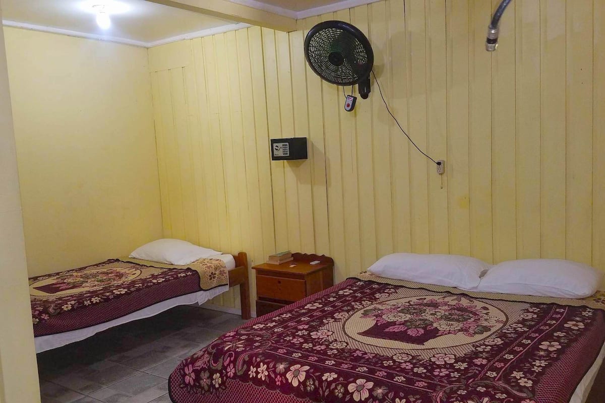 A room with two beds, each with a floral bedspread, a small wooden nightstand, a wall-mounted fan, and a telephone.