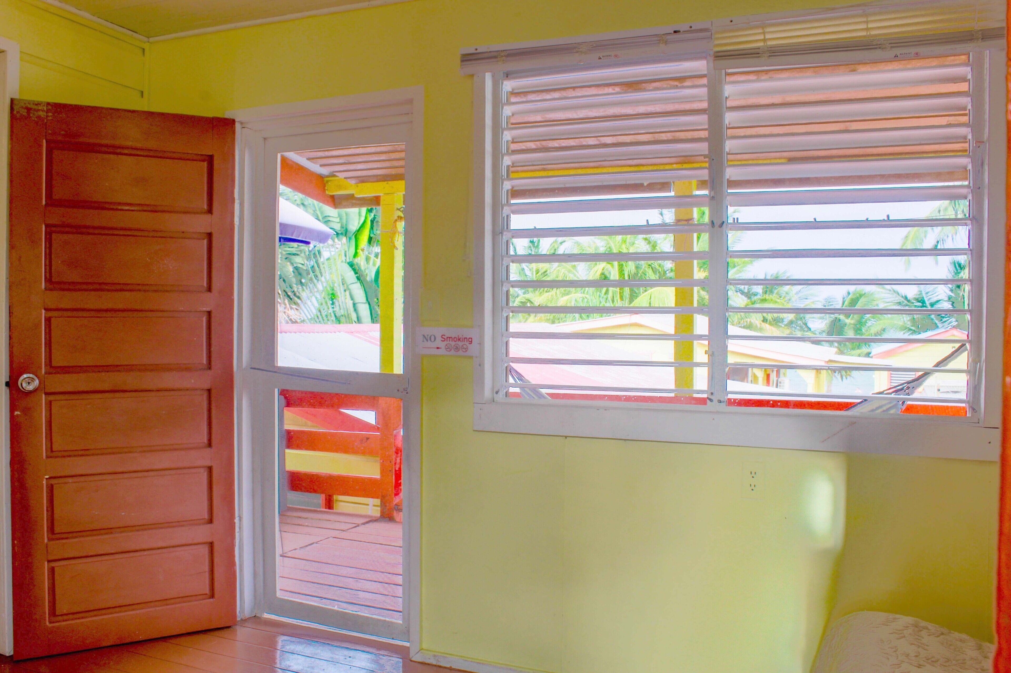 A room with a wooden door, a window with horizontal blinds, and a view of a colorful outdoor area.
