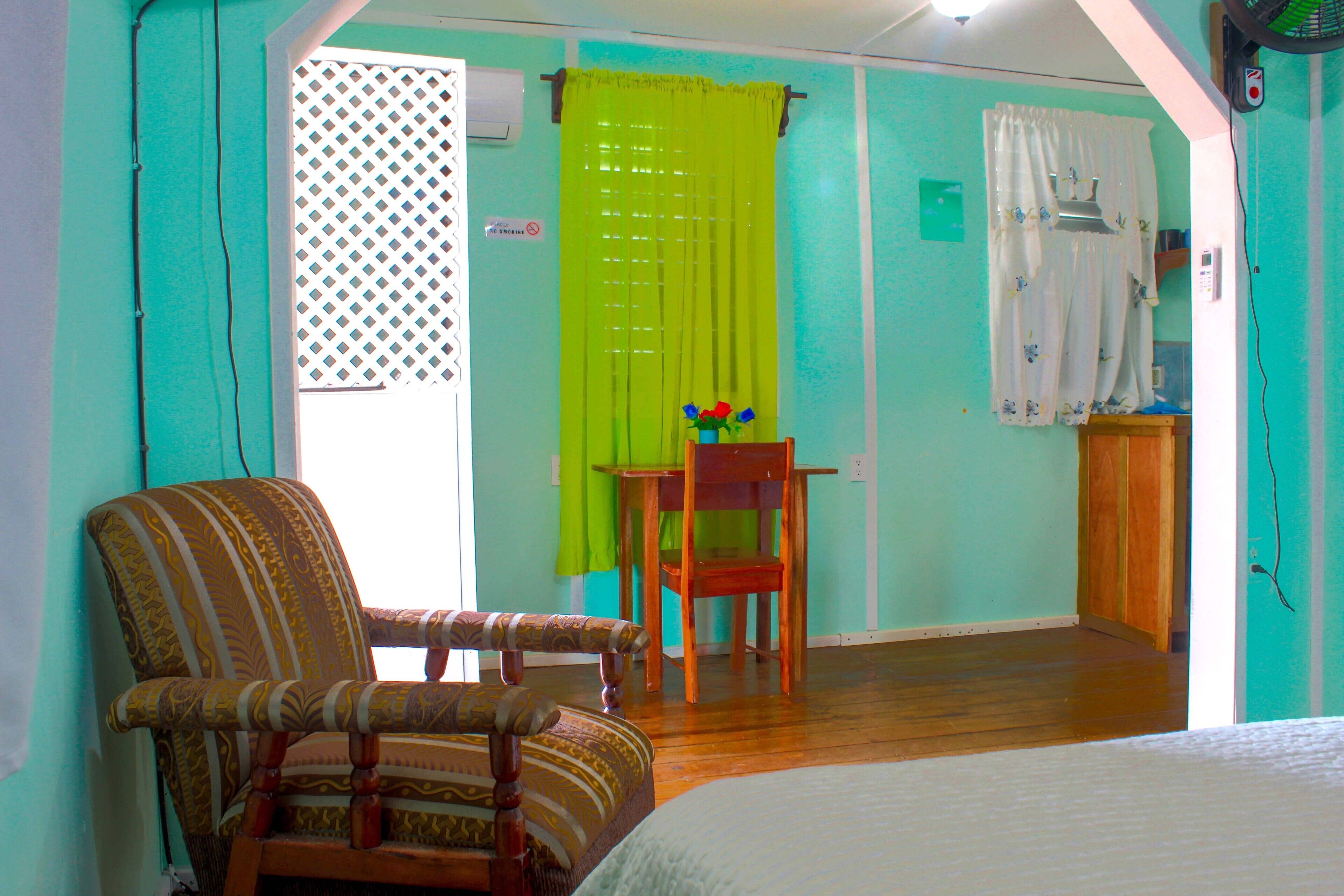 A room with a bed, a wooden chair, a small table with a vase of flowers, and a window with curtains.