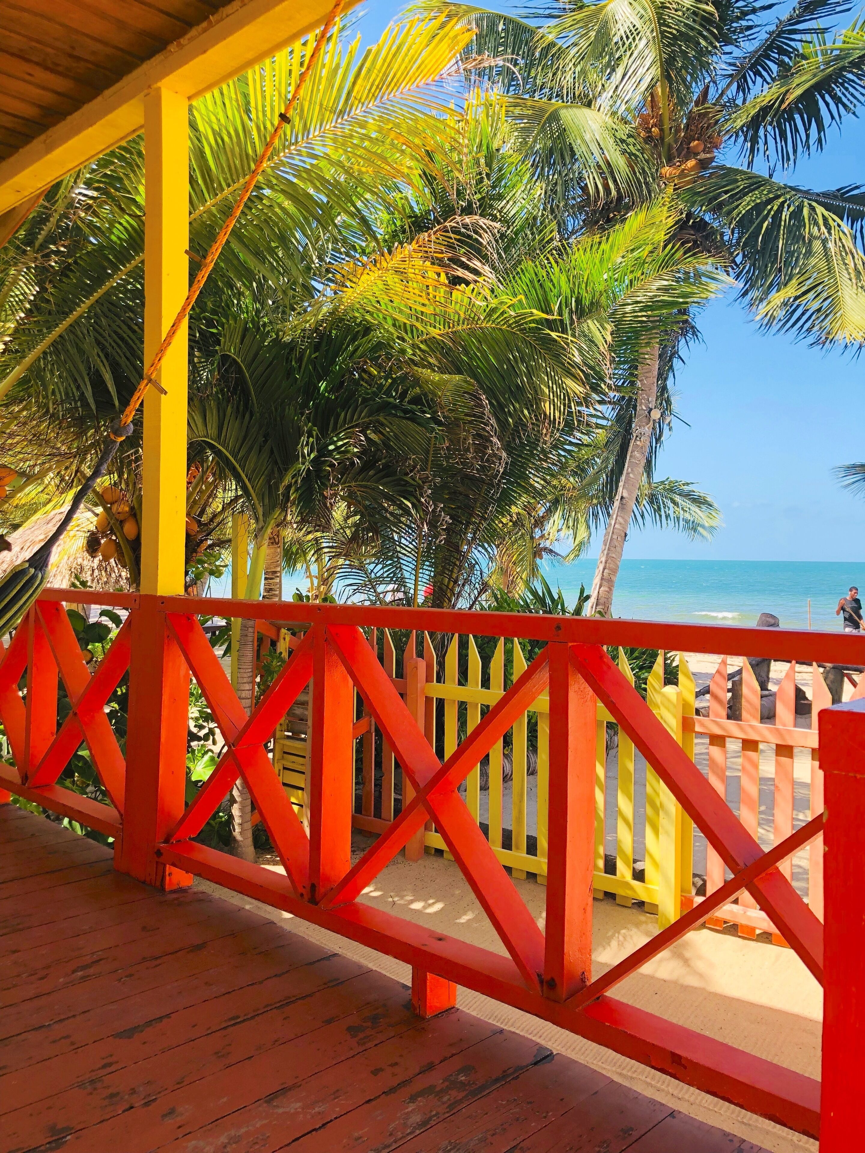 A wooden deck with a red railing overlooking a beach with palm trees and a clear blue sky.