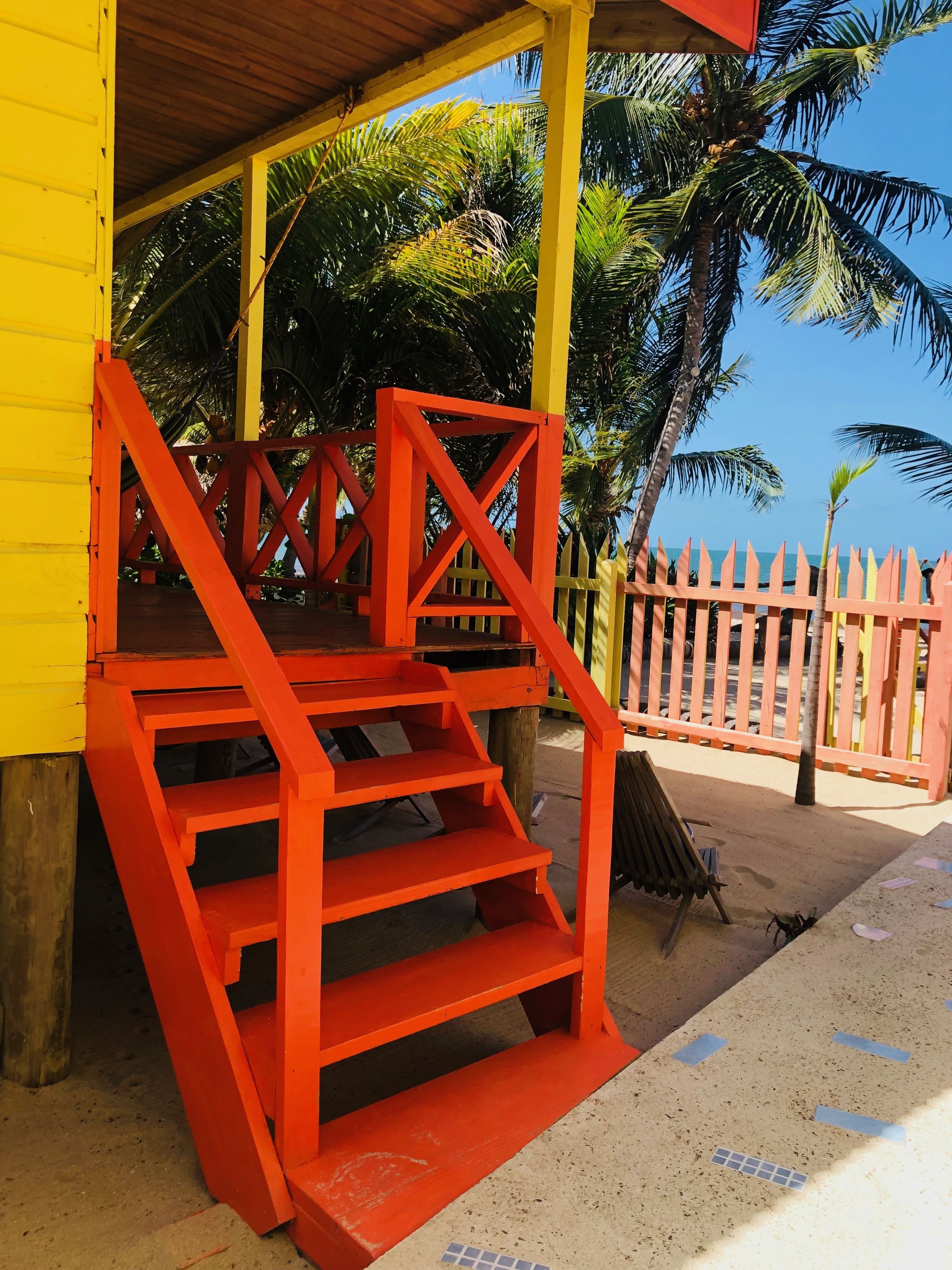 A staircase with red railings leading to a yellow building, surrounded by palm trees and a wooden fence.