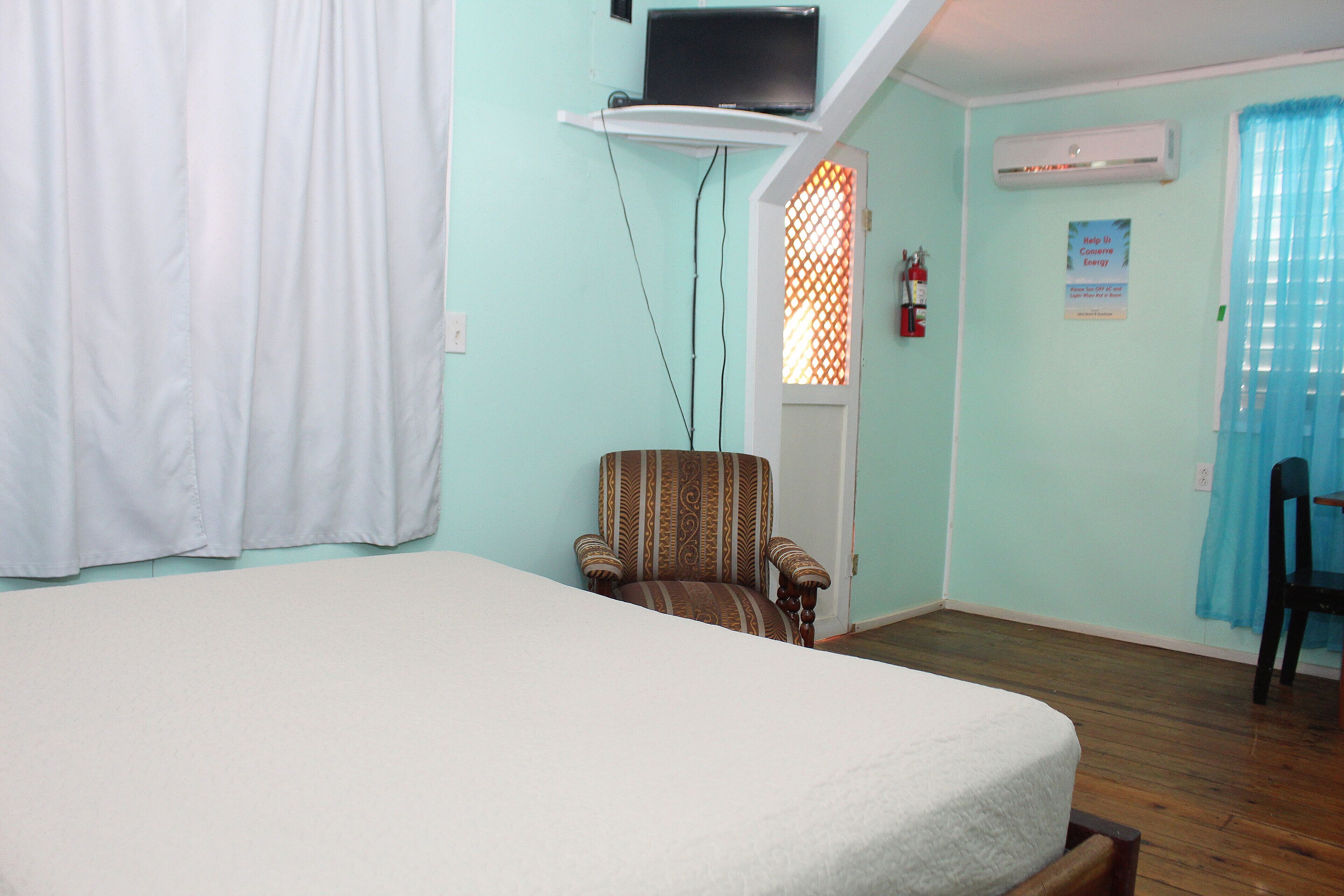 A room with a bed, a chair, a television on a shelf, an air conditioner, and a window with a curtain.