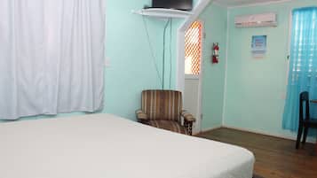 Una habitación con cama, una silla, televisor en estante, aire acondicionado y ventana con cortina.