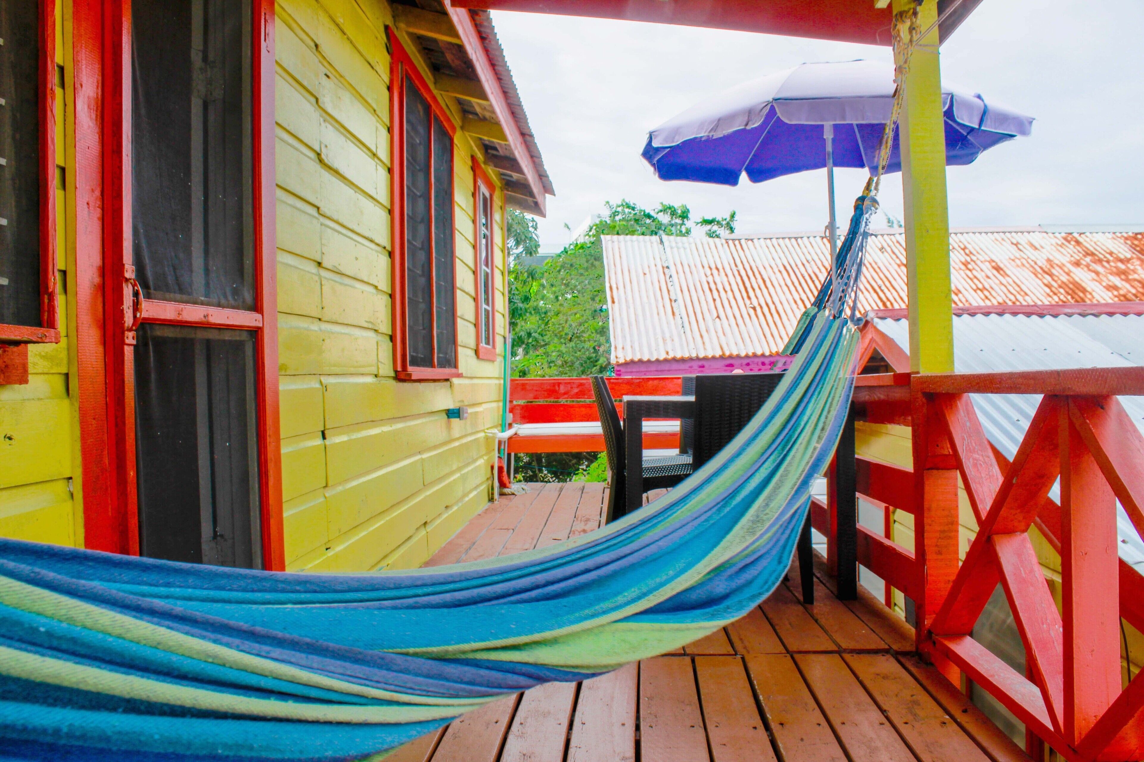 Eine Hängematte auf einer Holzdielen-Terrasse, im Hintergrund ein gelbes Gebäude und ein rotes Geländer.