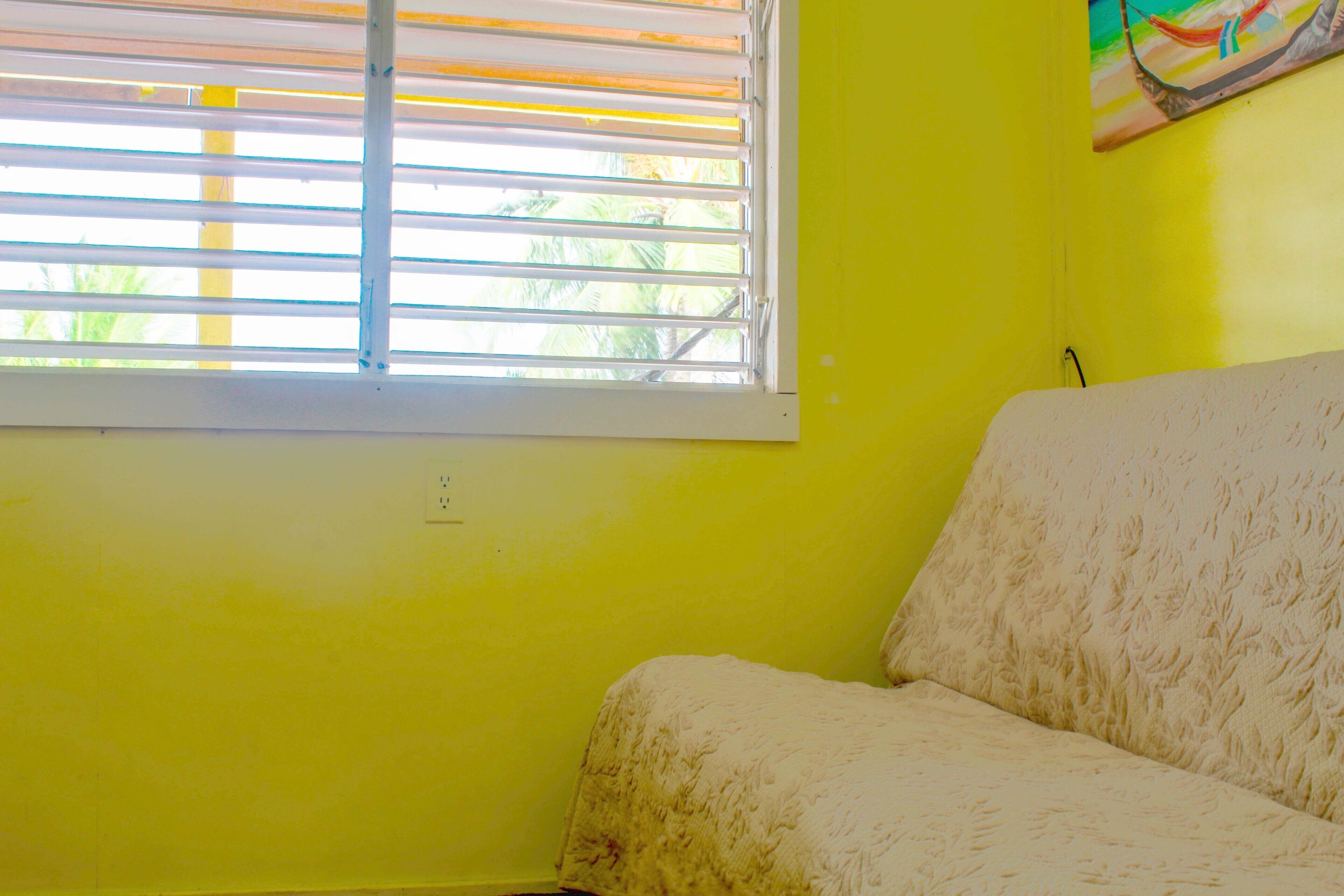 Quarto com parede amarela, janela com persianas e sofá branco.