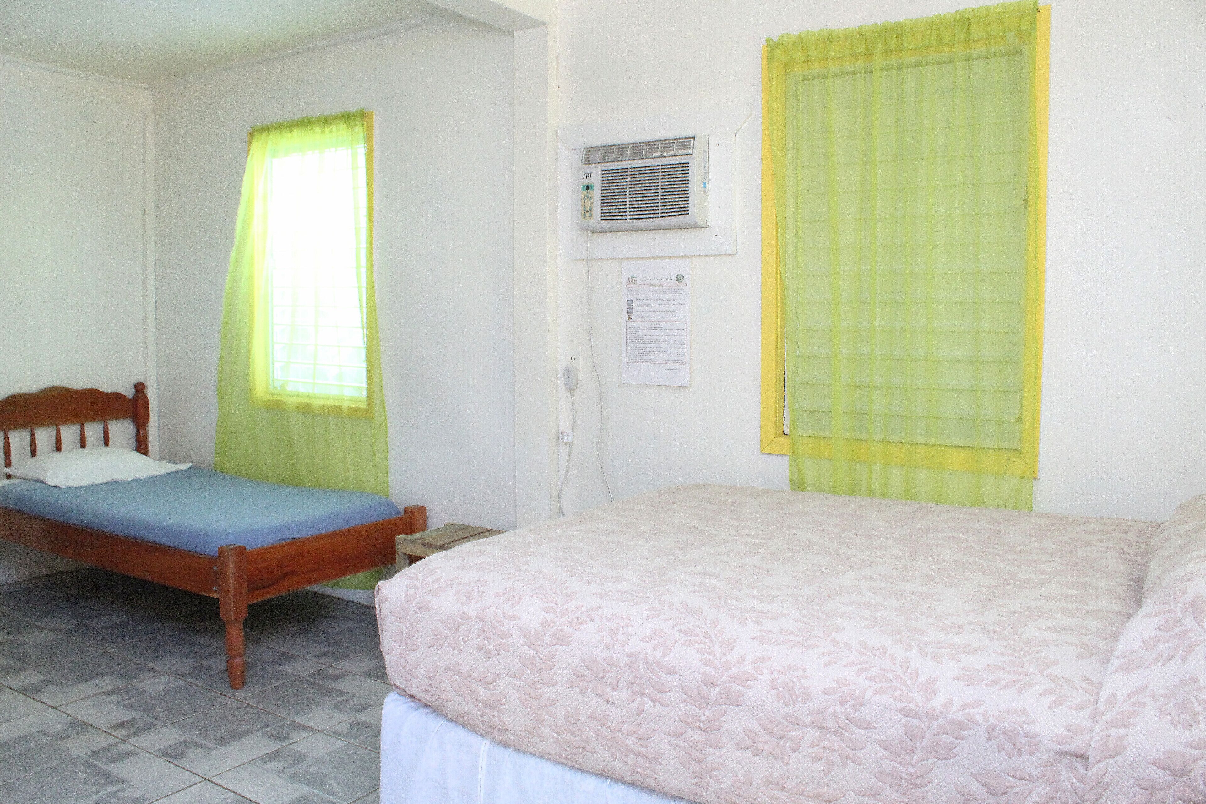 A room with two beds, one with a wooden frame and the other with a metal frame. There is a window with a yellow curtain, an air conditioning unit on the wall, and a small table beside the wooden bed.