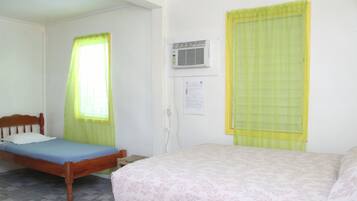 A room with two beds, one with a wooden frame and the other with a metal frame. There is a window with a yellow curtain, an air conditioning unit on the wall, and a small table beside the wooden bed.