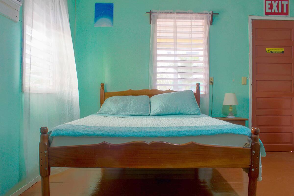 A bed with a wooden frame and a blue bedspread in a room with teal walls, a window with white curtains, and an exit sign on the door.