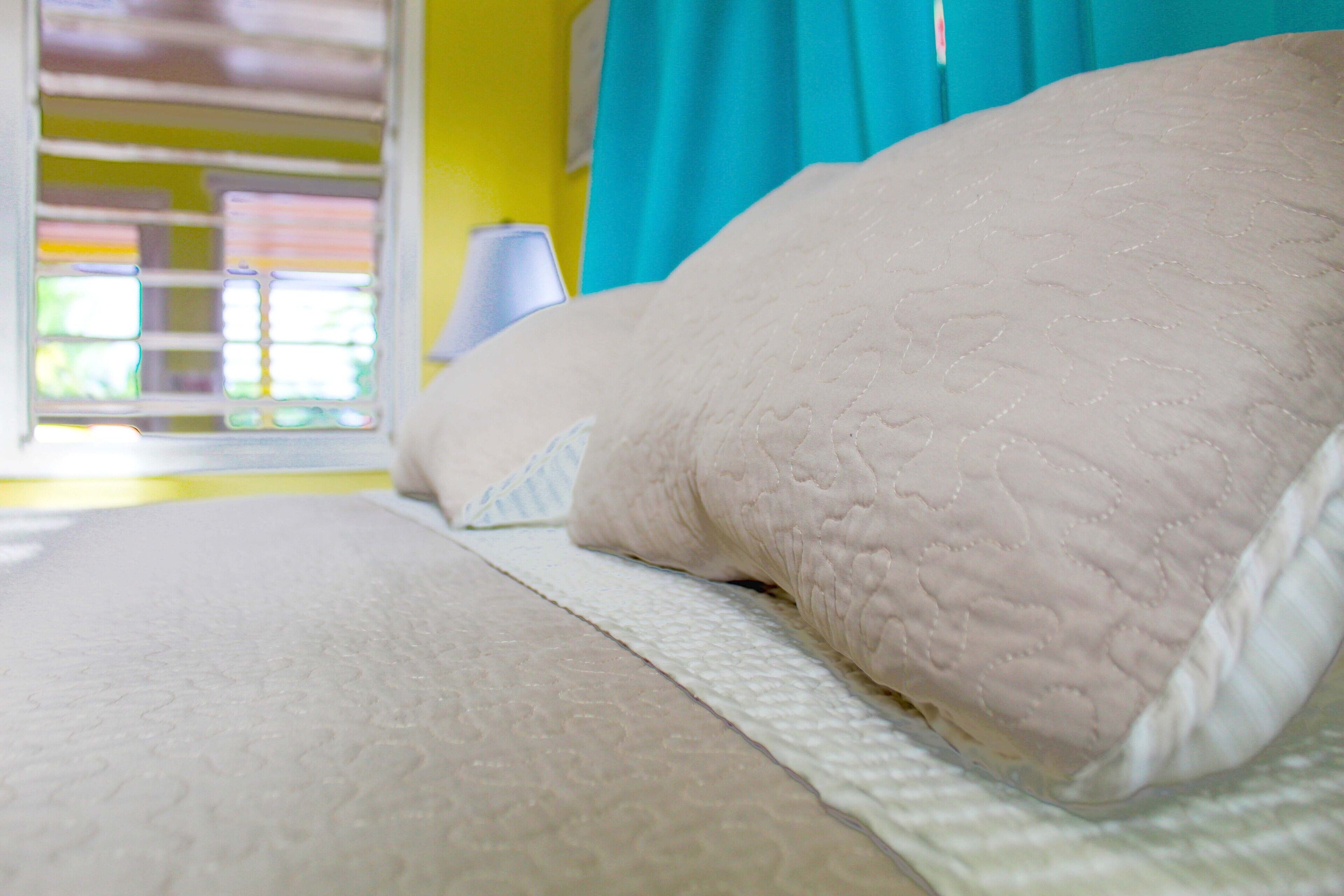 Una cama con colcha blanca y almohada, una ventana con persianas y cortina azul.