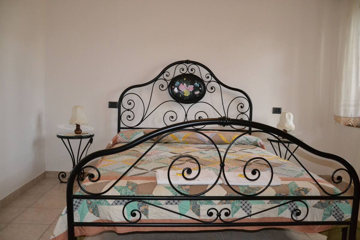 A bed with a metal frame and a quilt, a small table with a lamp, and a wall-mounted decoration.