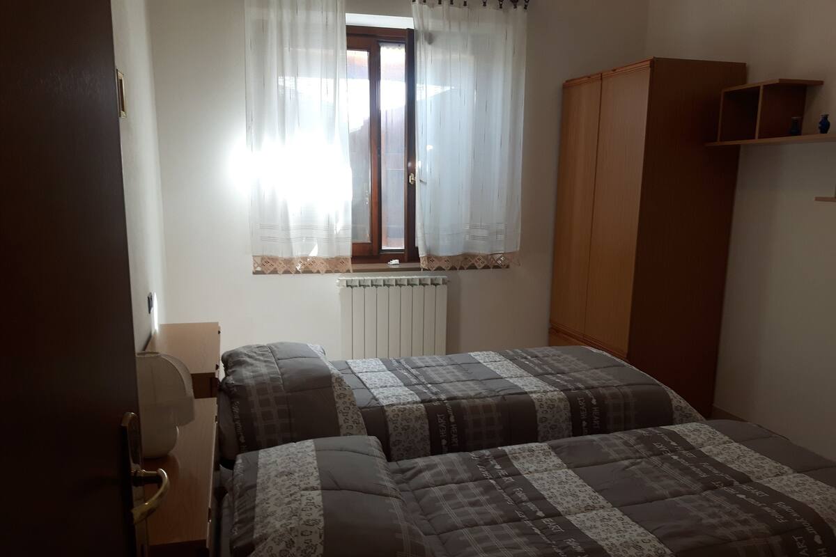 A bedroom with two beds, a radiator, a wardrobe, and a window with sheer curtains.