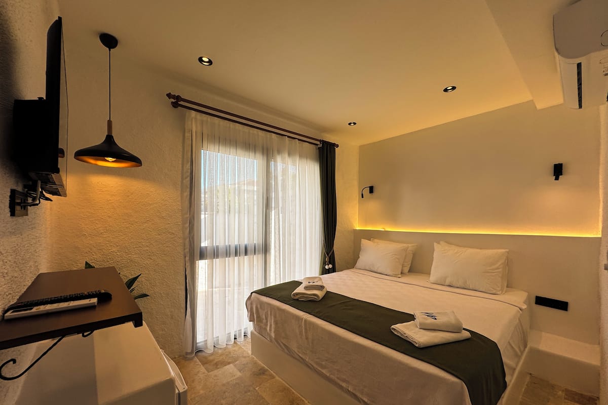 Habitación de hotel moderna con una cama bien hecha, un escritorio con una computadora portátil, un televisor de pantalla plana en la pared y un ventanal con cortinas ligeras.
