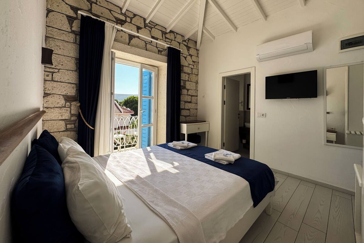 Un dormitorio con pared de piedra, balcón con contraventanas azules, cama con ropa de cama blanca y azul, televisor montado en la pared y unidad de aire acondicionado blanca.
