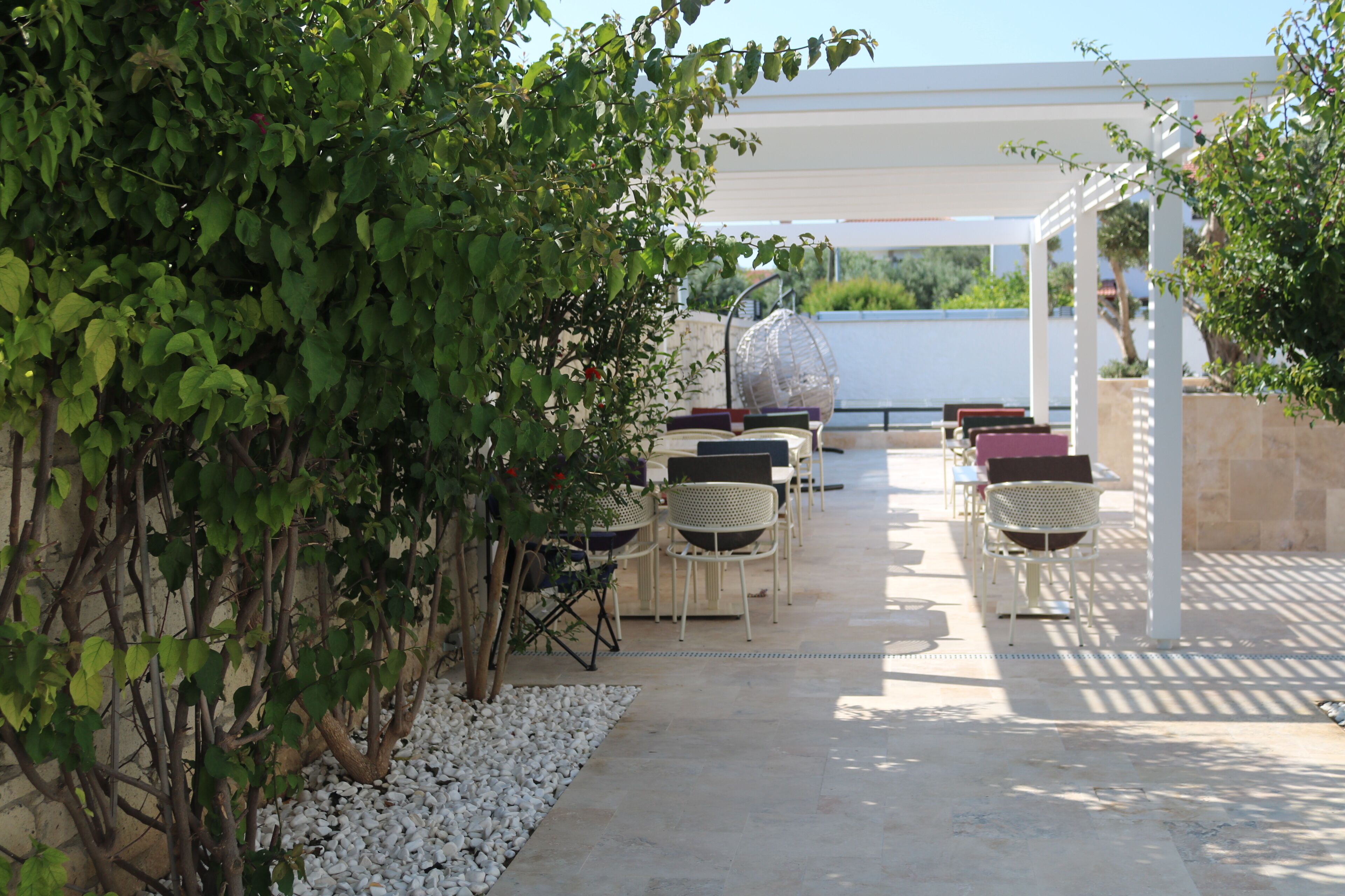An outdoor patio with white chairs and tables, surrounded by greenery and a pergola structure.