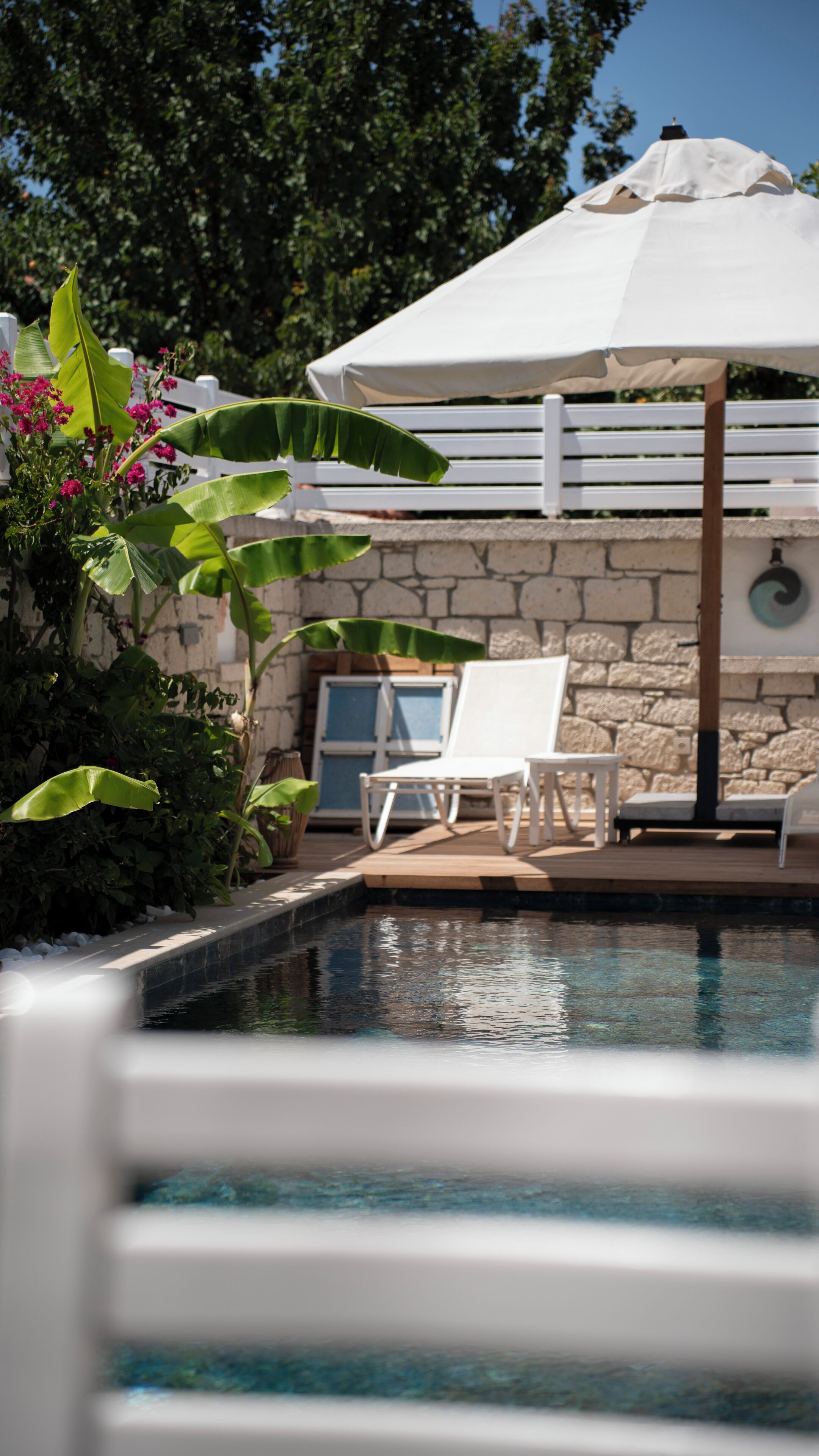 A pool area with a white umbrella, white lounge chairs, and a stone wall in the background.