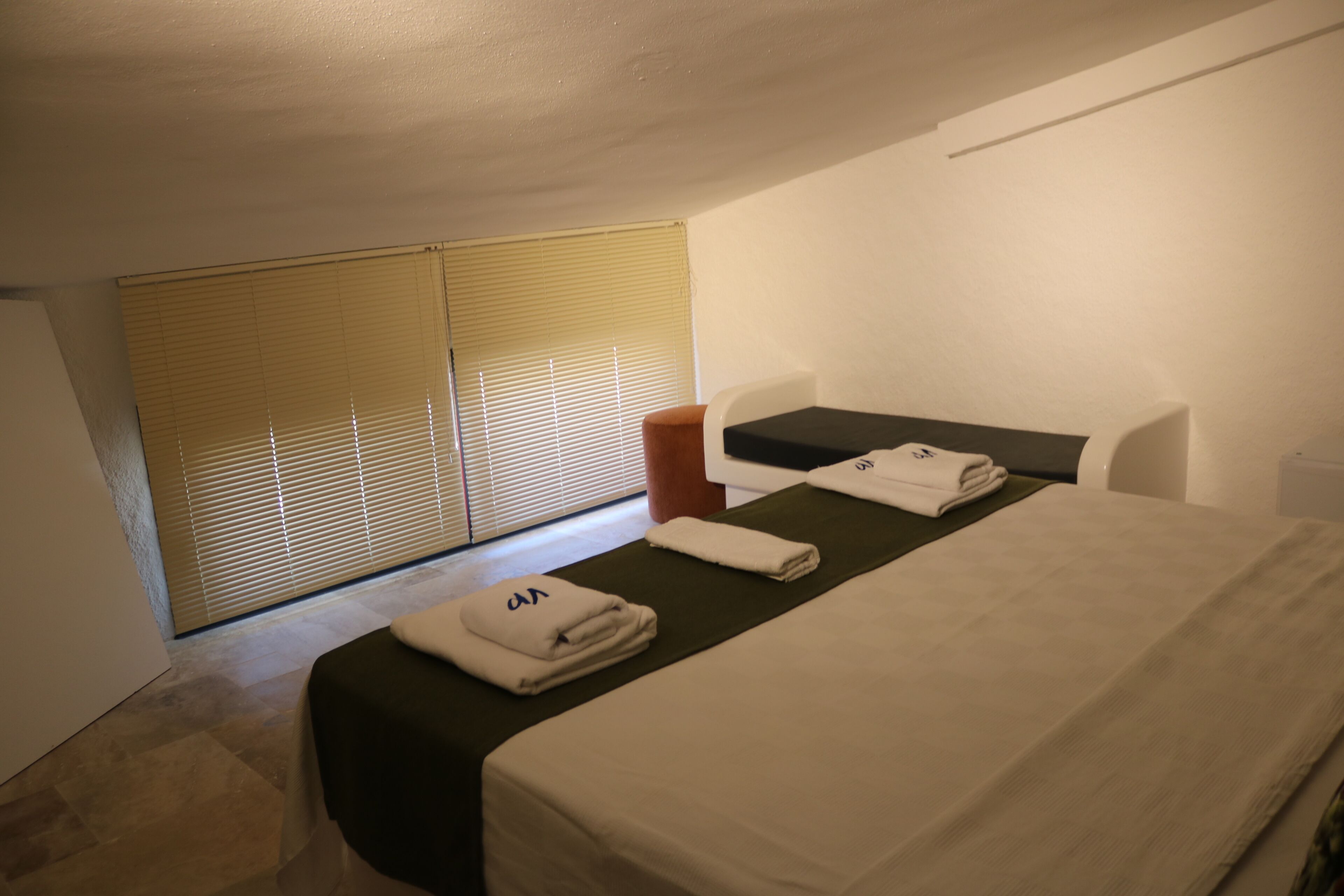 A hotel room with a bed, a small table, a chair, a window with blinds, and a wall-mounted TV.