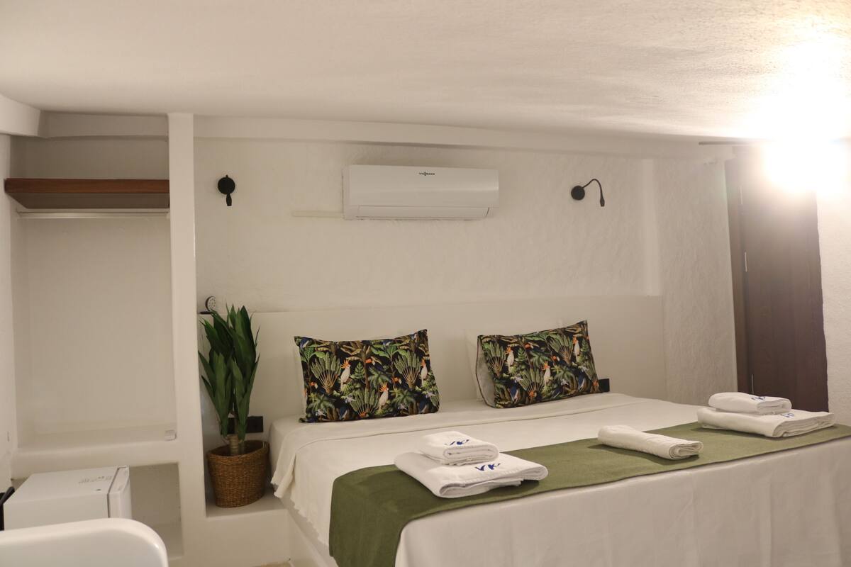 Habitación de hotel con cama, almohadas, colcha verde, planta en maceta, aire acondicionado montado en la pared y una mesita con toallas.
