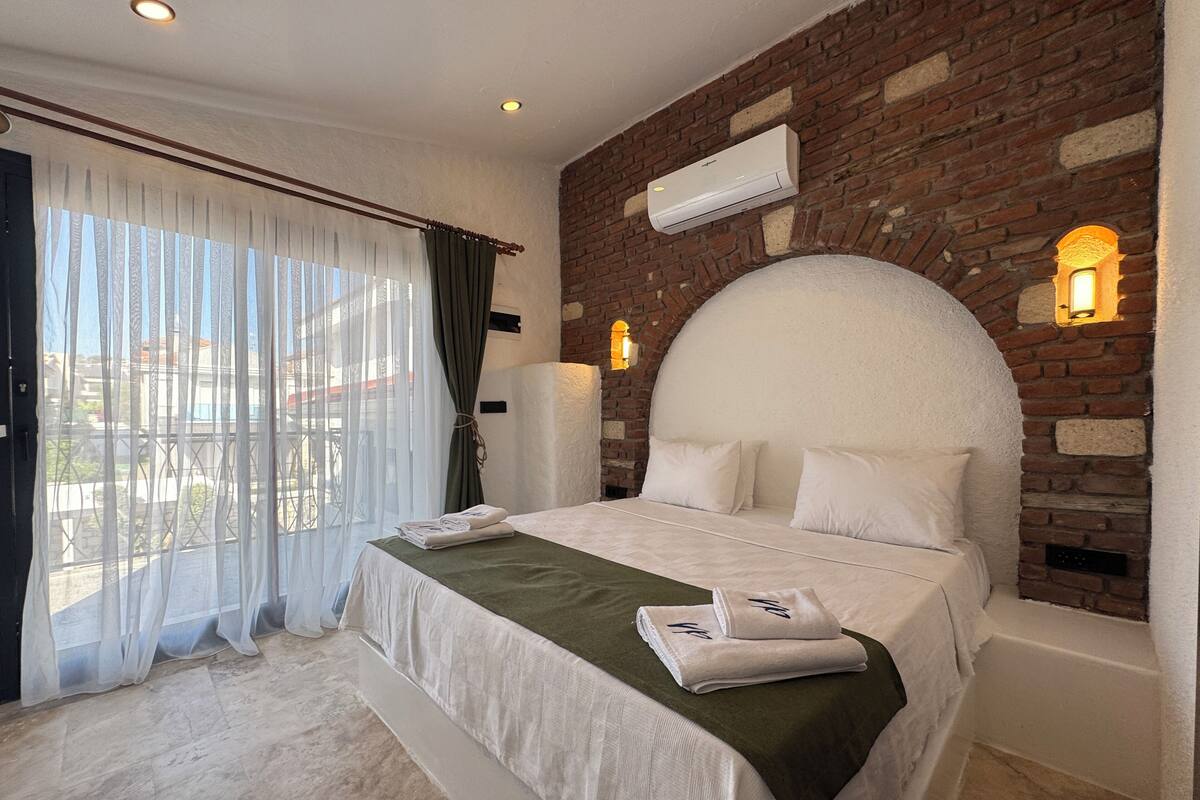 Habitación de hotel con pared de ladrillo, una cama grande con ropa blanca, un plaid verde y vistas a través de cortinas translúcidas.