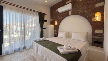 Une chambre d’hôtel avec un mur en briques, un grand lit recouvert de draps blancs, un plaid vert et une vue à travers des rideaux transparents.