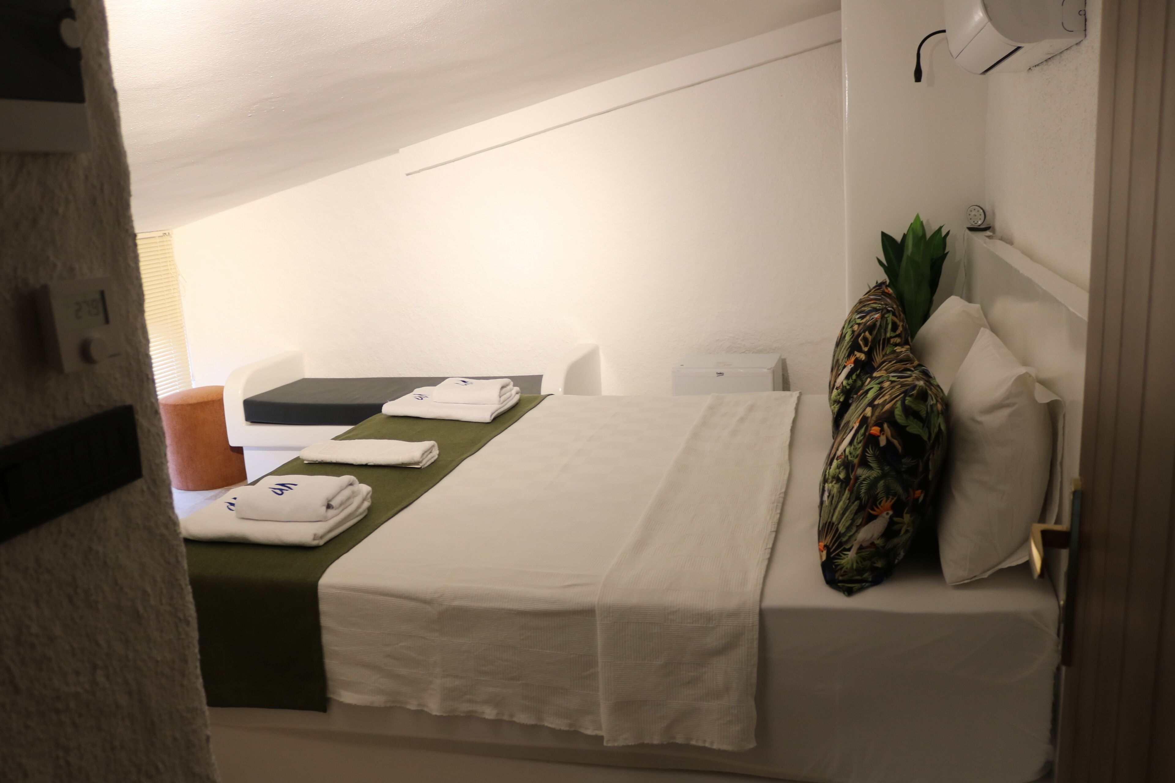 A hotel room with a bed, a small table, a chair, a plant, and a wall-mounted heater.