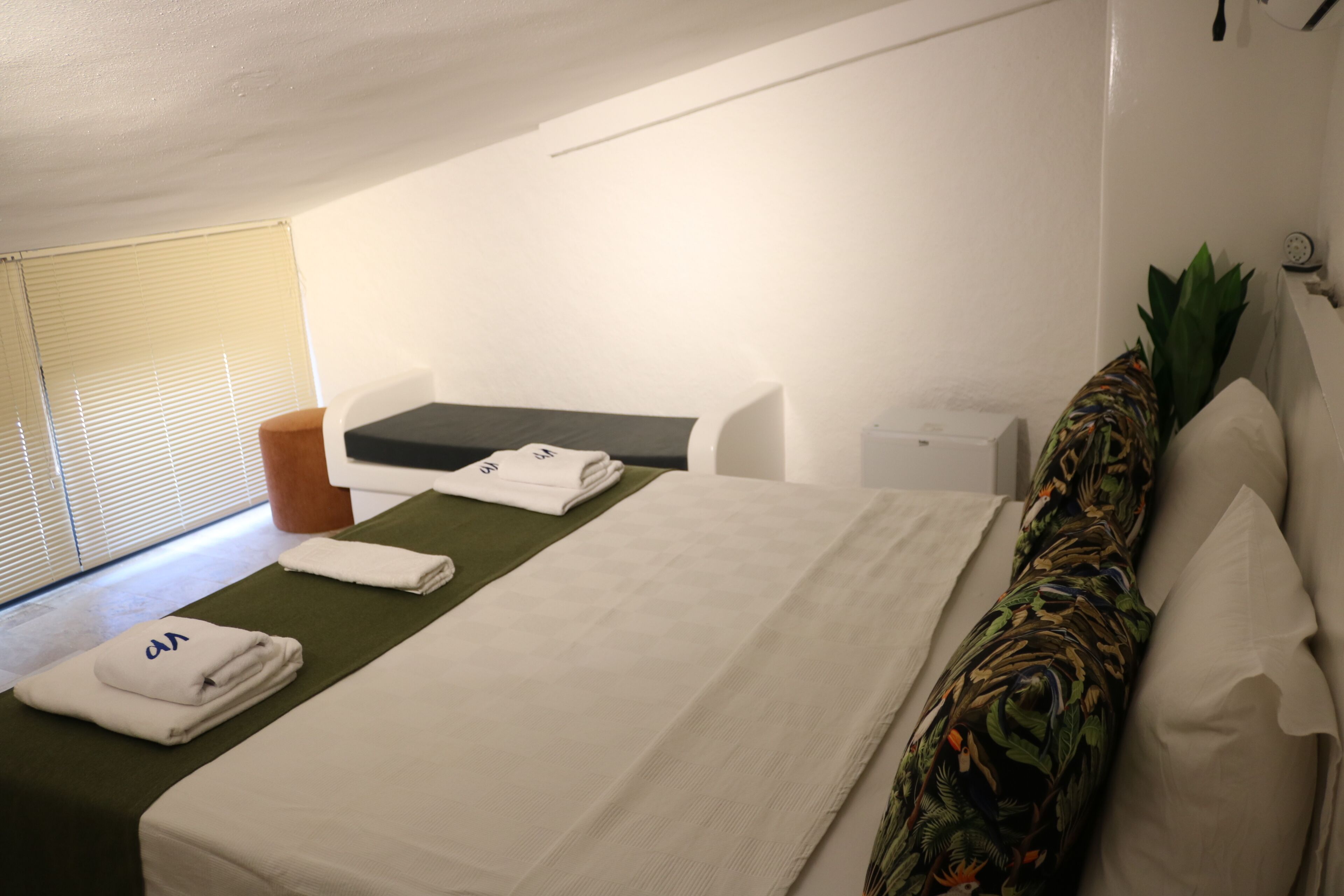 Une chambre d’hôtel avec un lit, un petit bureau, une chaise, une fenêtre avec des stores et une plante.