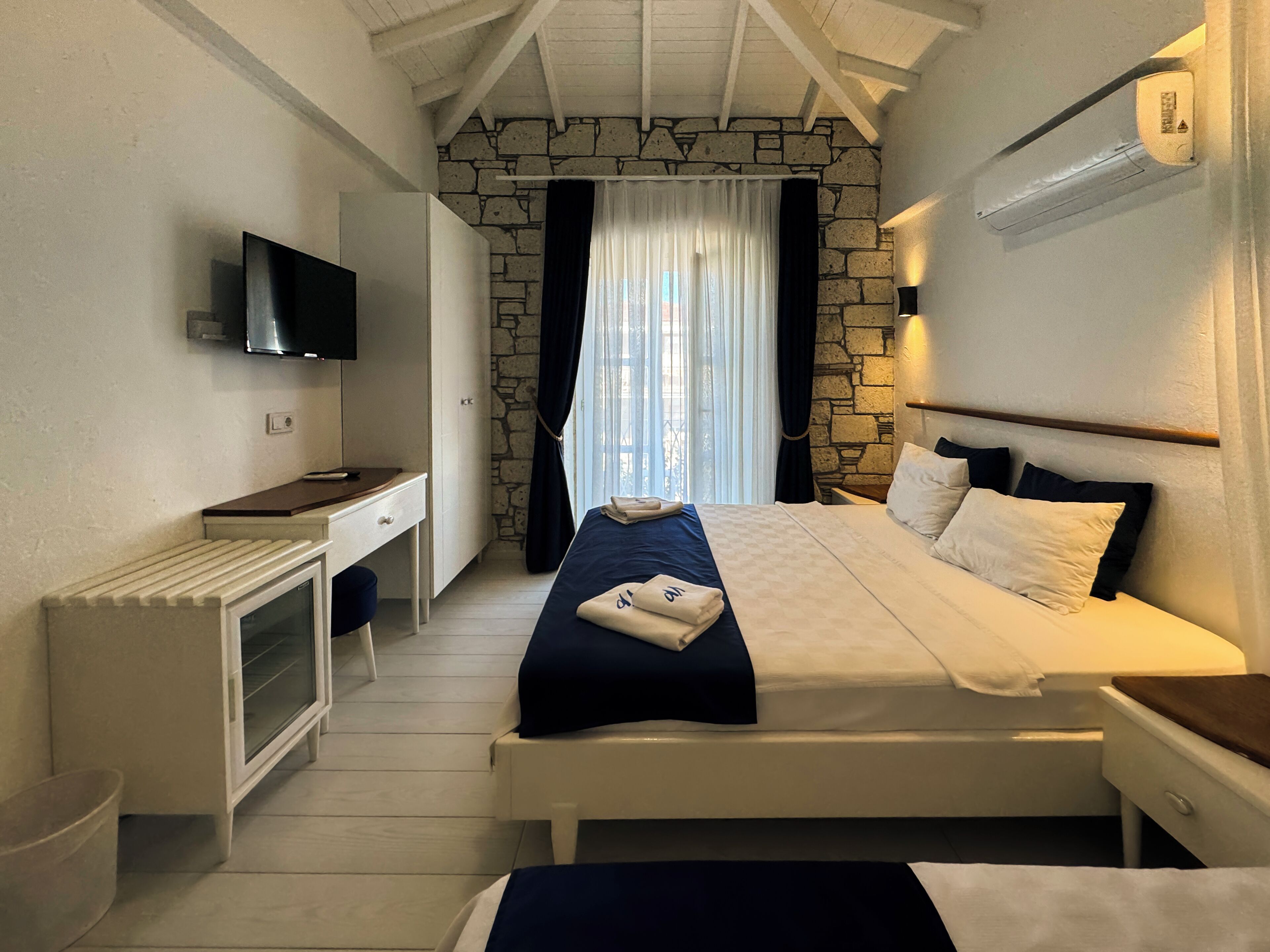 Une chambre avec un mur en pierre, un lit avec une literie blanche et bleu foncé, un bureau avec un téléviseur à écran plat et une fenêtre avec des rideaux.