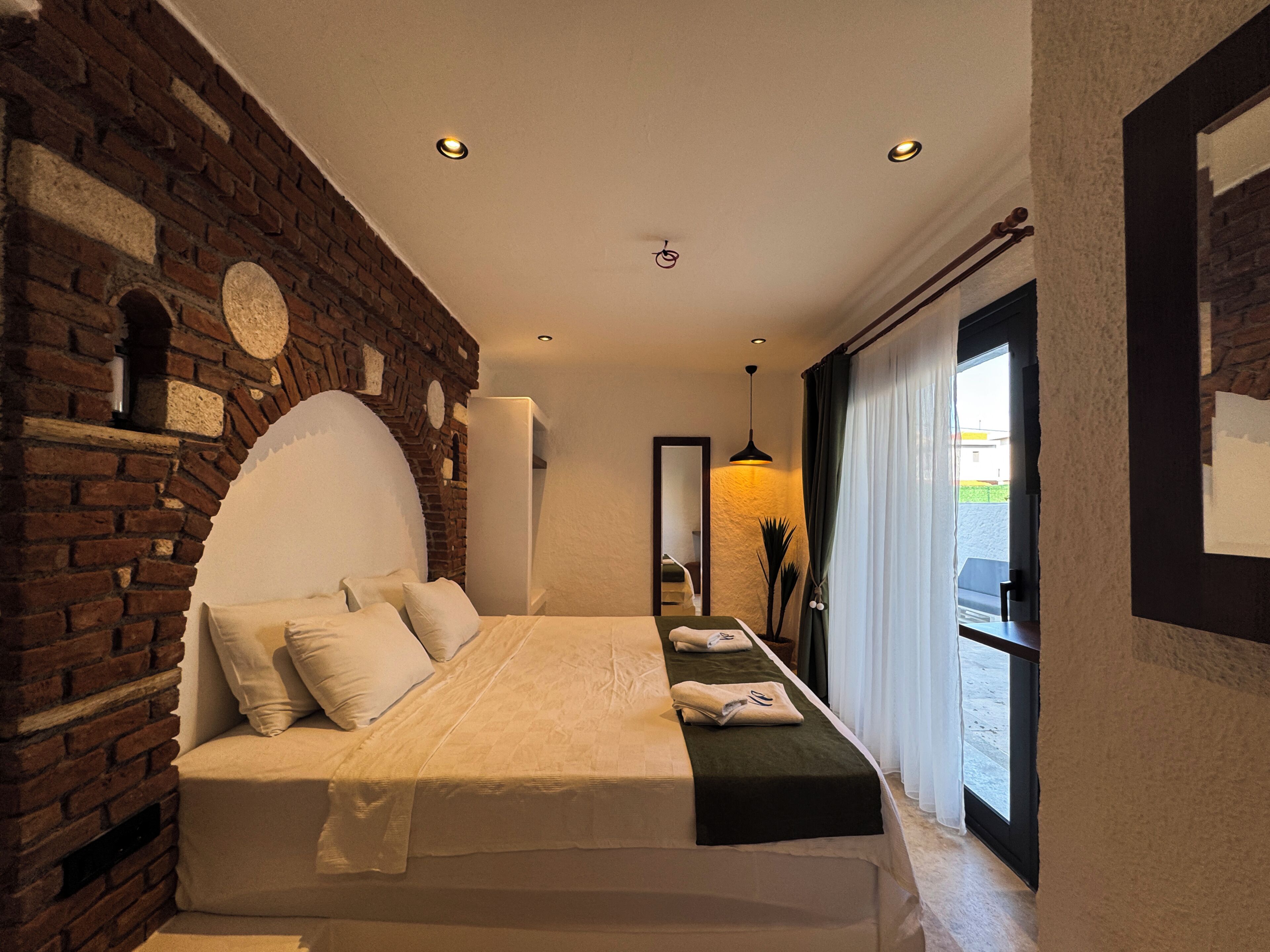 A bedroom with a brick wall, a bed with pillows, a bedside table, and a view through a window.