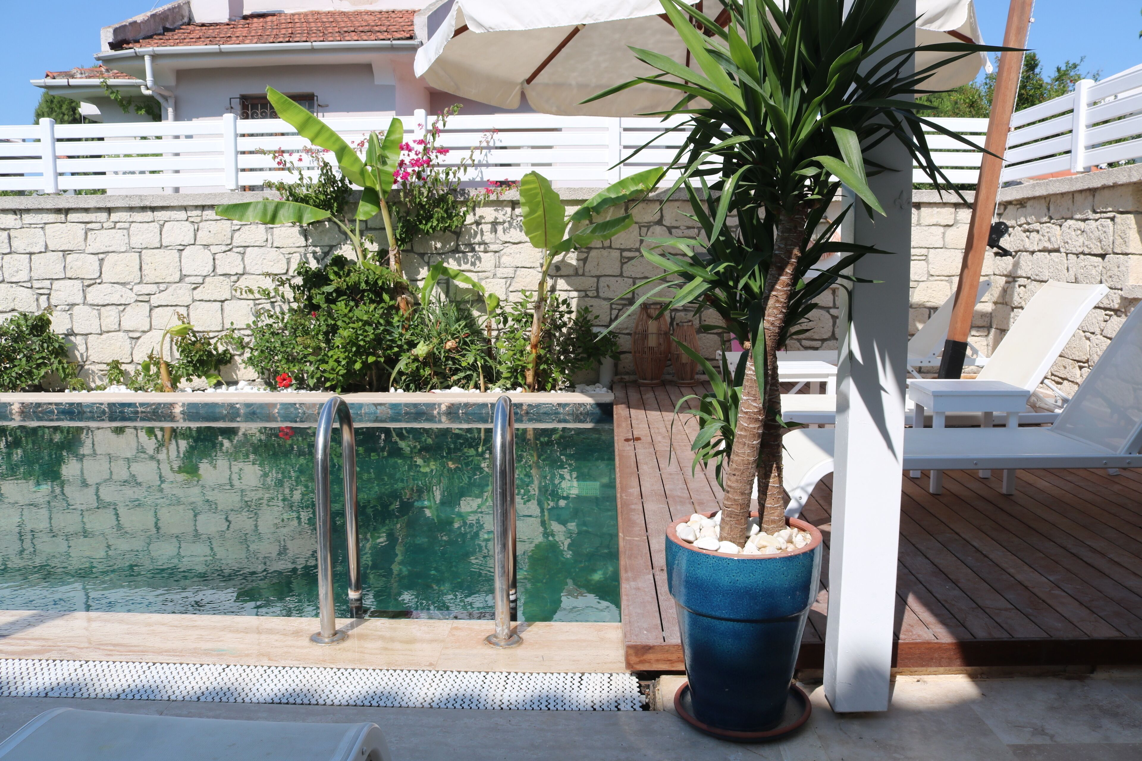 Zona de piscina con muro de piedra, terraza de madera y sillas reclinables blancas.