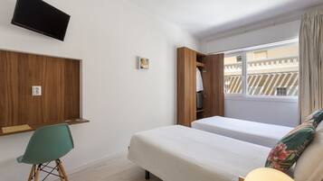 A hotel room with two beds, a TV mounted on the wall, a desk with a chair, and a window with a view of buildings.