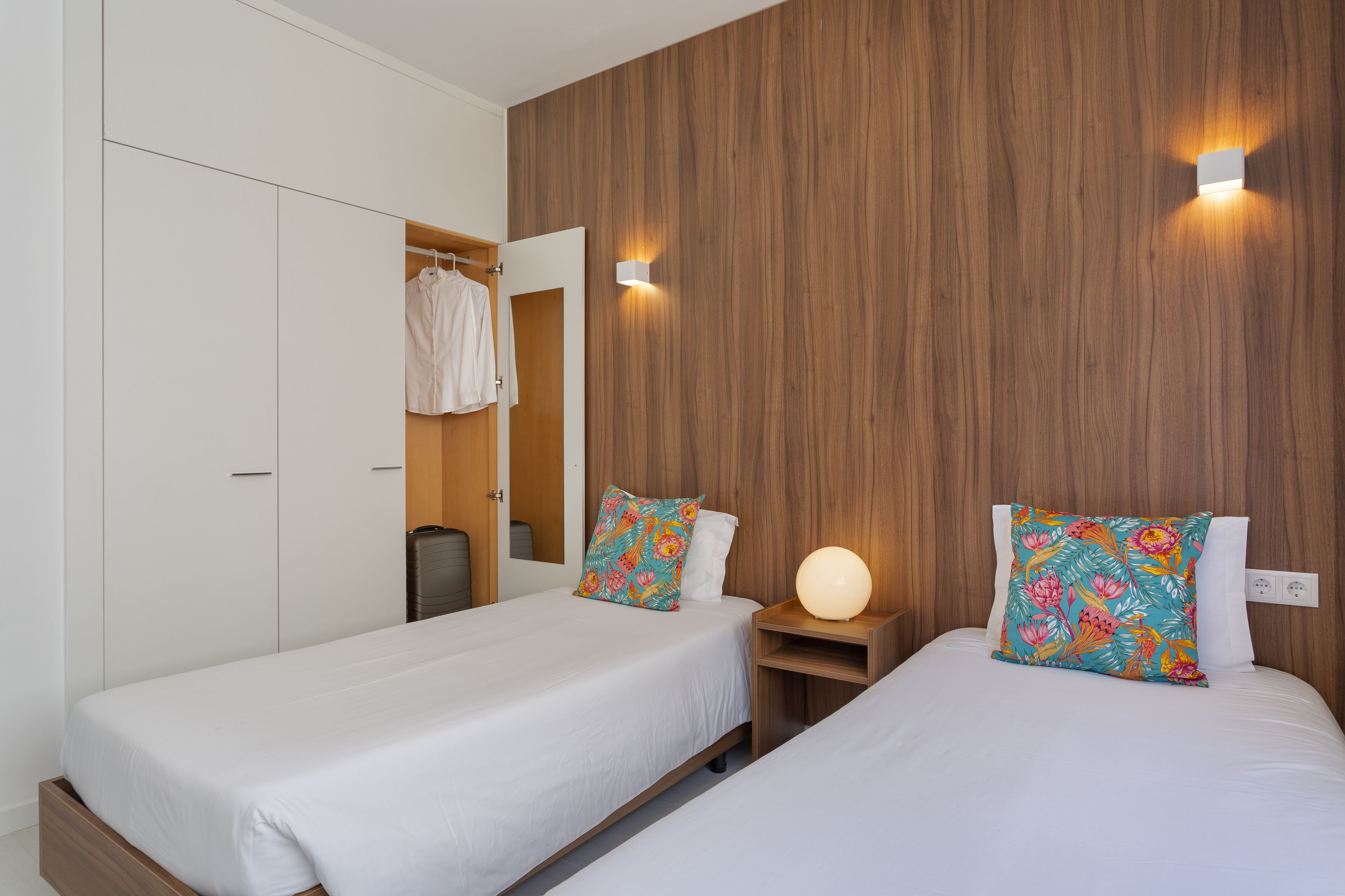 A bedroom with two beds, wooden headboard, white bedding, a nightstand with a lamp, and a wardrobe.