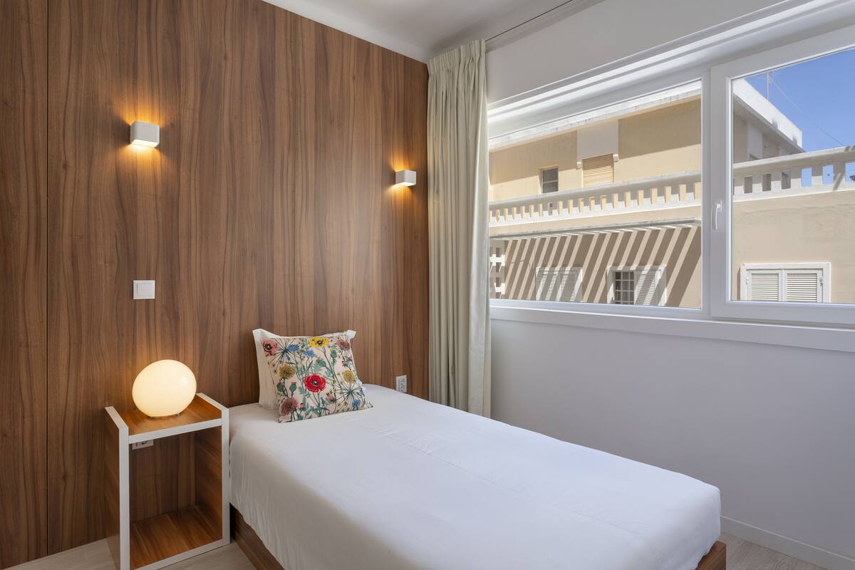 Quarto com parede de madeira, mesa de cabeceira com luminária, cama com travesseiro floral e janela com vista para uma varanda.