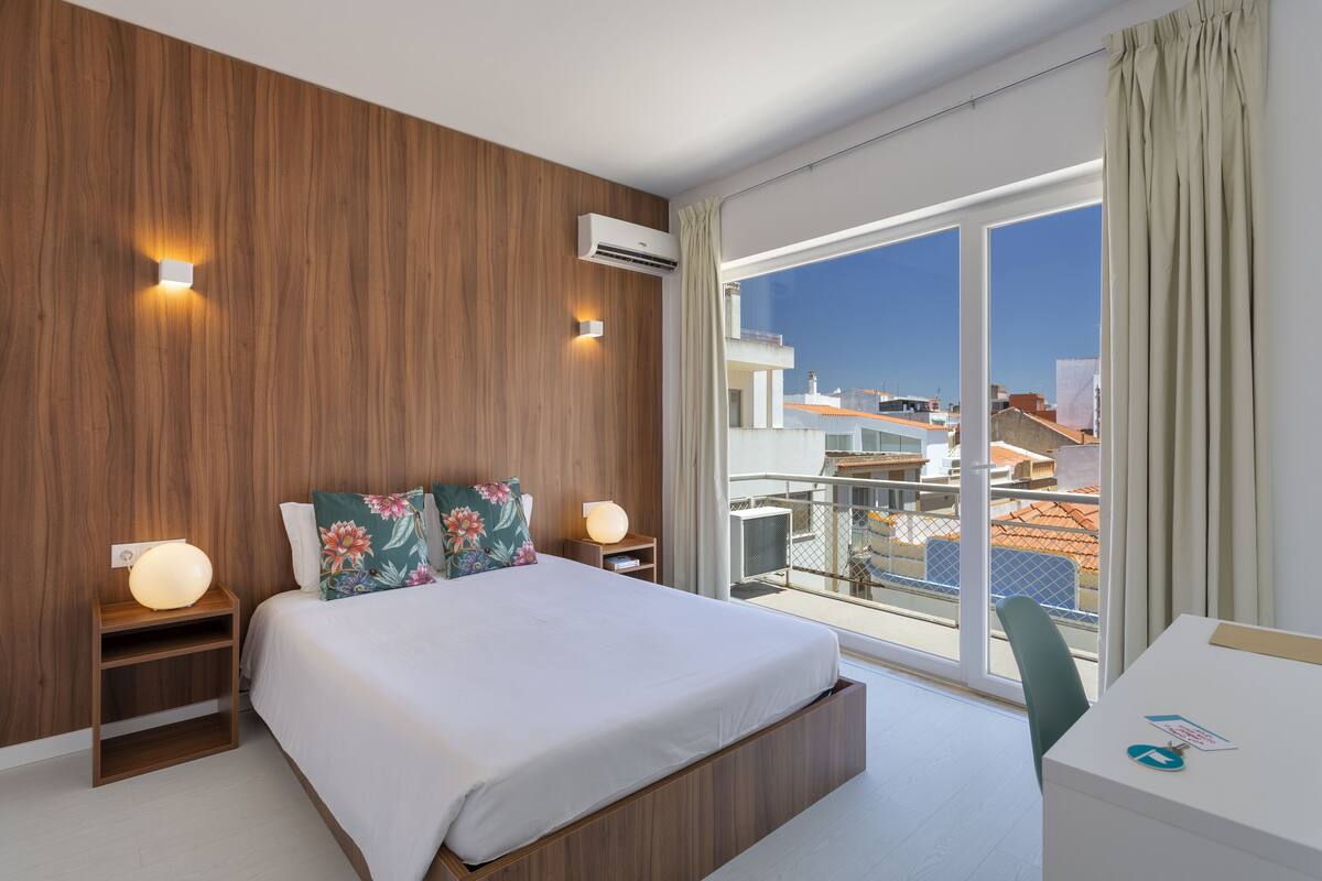 Quarto de hotel moderno com uma cama grande, painéis de madeira, uma escrivaninha e uma varanda com vista para a cidade.