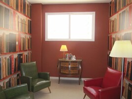 Una habitación con un ventanal grande, dos sillones, una lámpara y estanterías llenas de libros.