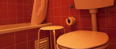 A bathroom with red tiled walls, a white toilet, a shower with a white curtain, and a small stool.
