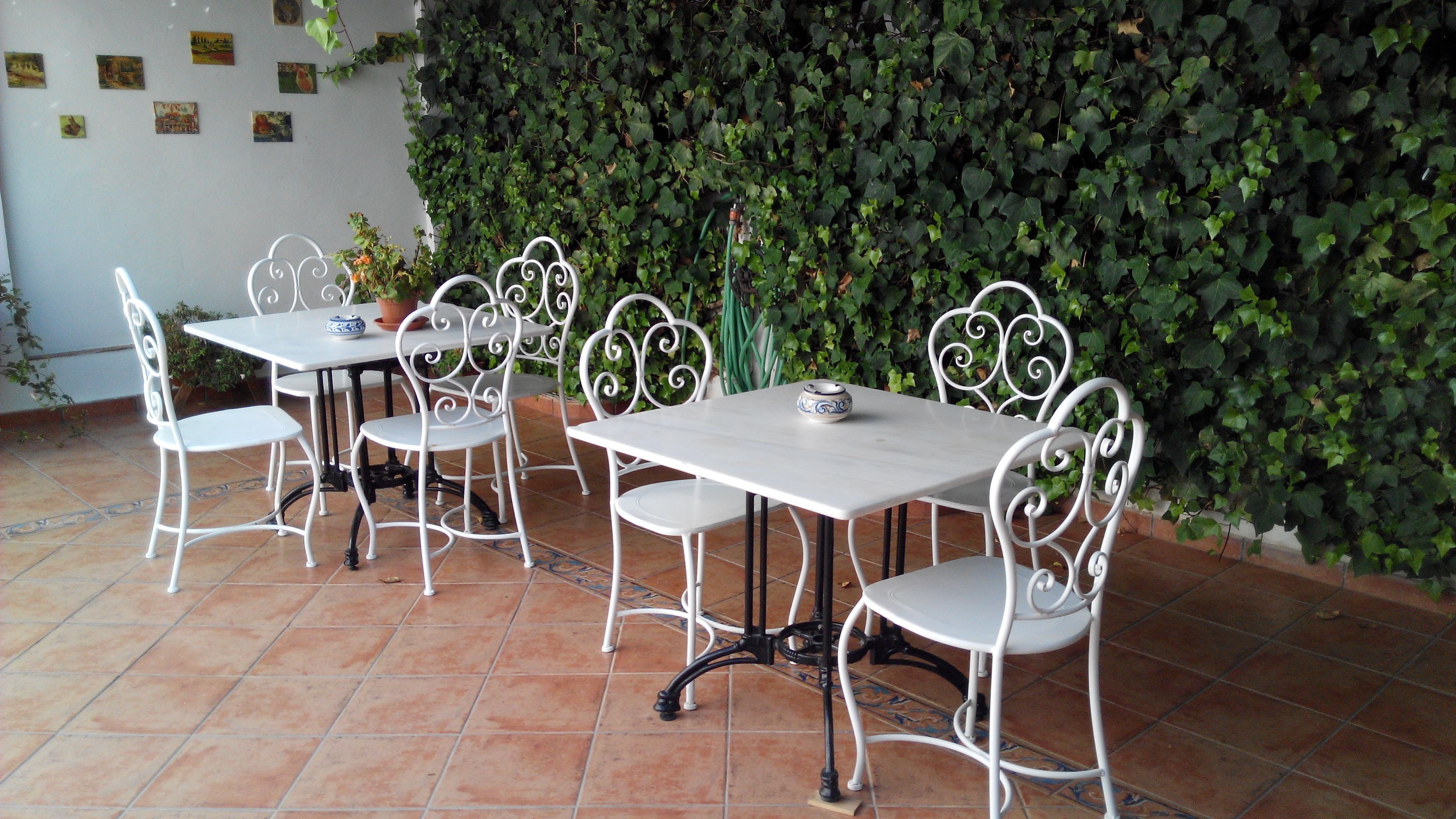 Un patio con mesas y sillas de metal blanco, rodeado por un muro cubierto de hiedra verde.
