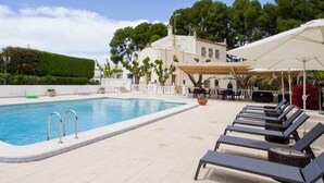 Área de piscina com espreguiçadeiras e guarda-sóis, cercada por um edifício e árvores.
