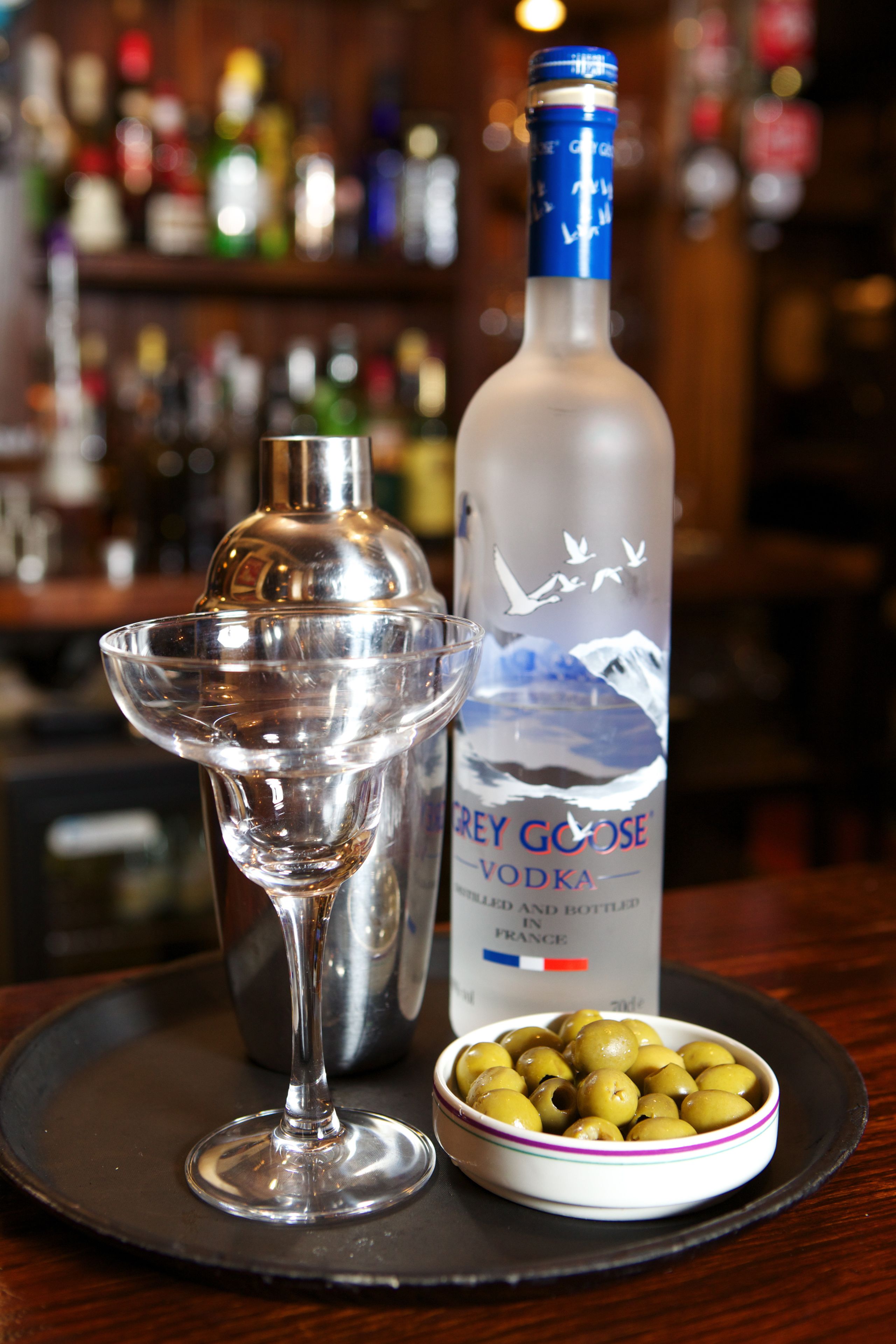 Un bar con una botella de Grey Goose Vodka, un coctelero y un bol de aceitunas sobre una bandeja.