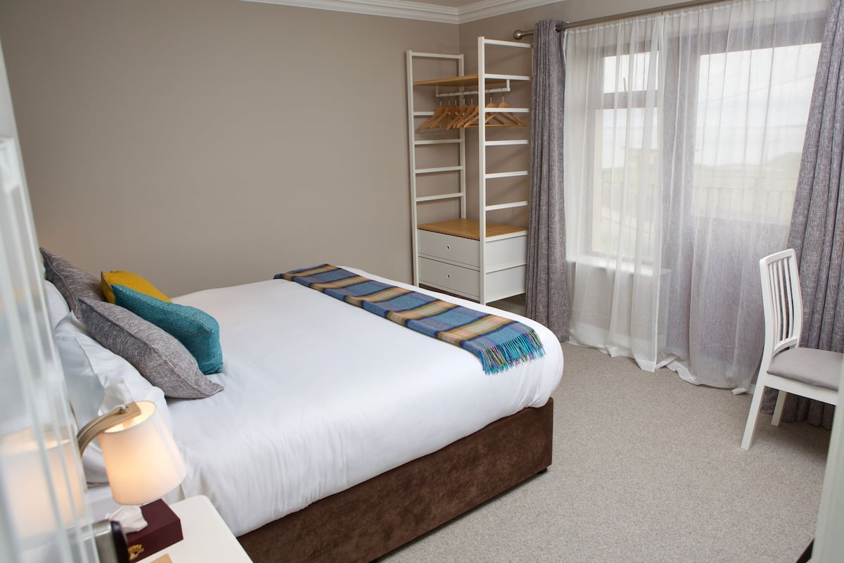 Ein ordentlich gemachtes Bett mit weißer Tagesdecke und Streifenmusterdecke, ein Nachttisch mit Lampe, ein weißer Stuhl, ein eingebauter Kleiderschrank mit Regalen und ein Fenster mit transparenten Vorhängen.