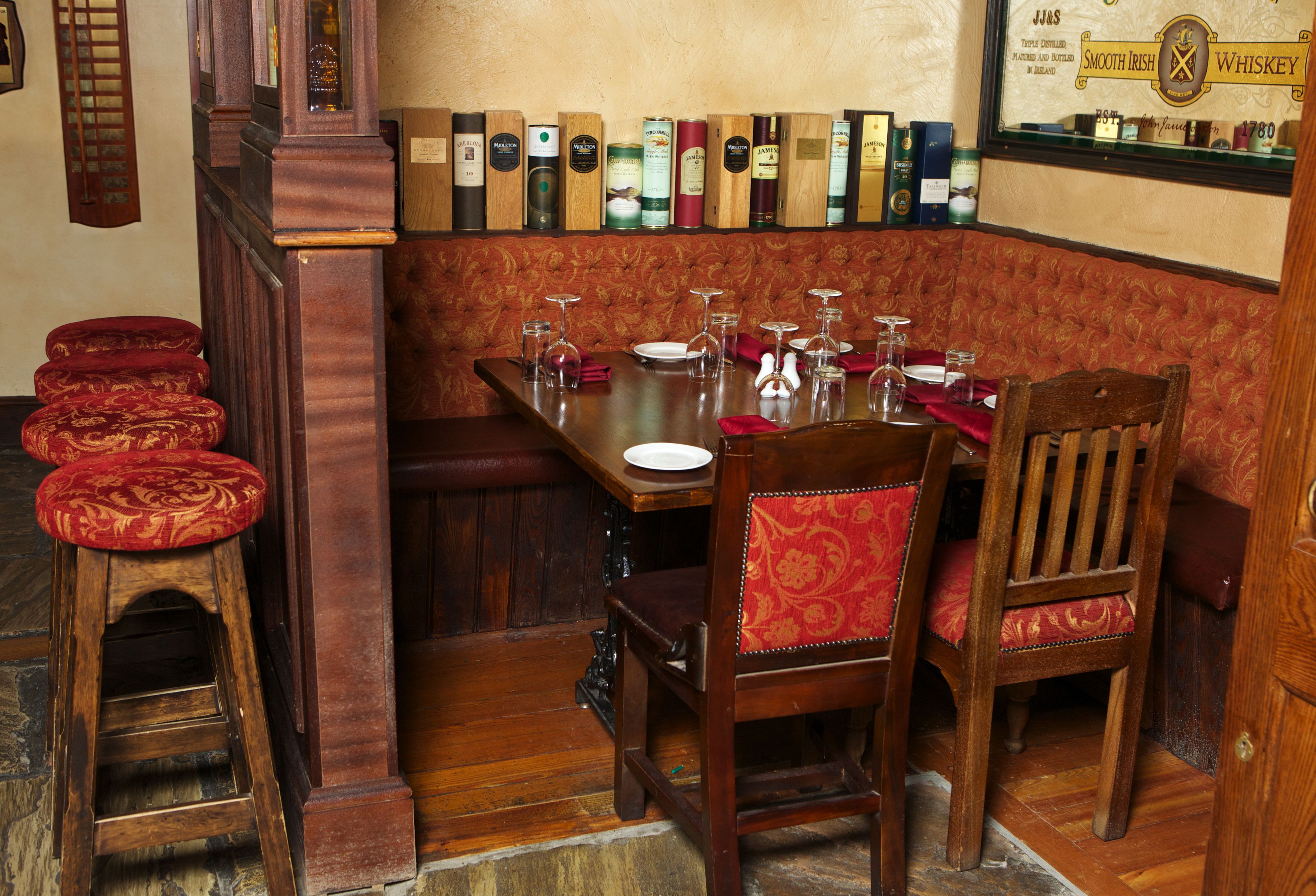 A traditional pub interior with a wooden table, red cushioned benches, and a selection of whiskey bottles on the wall.