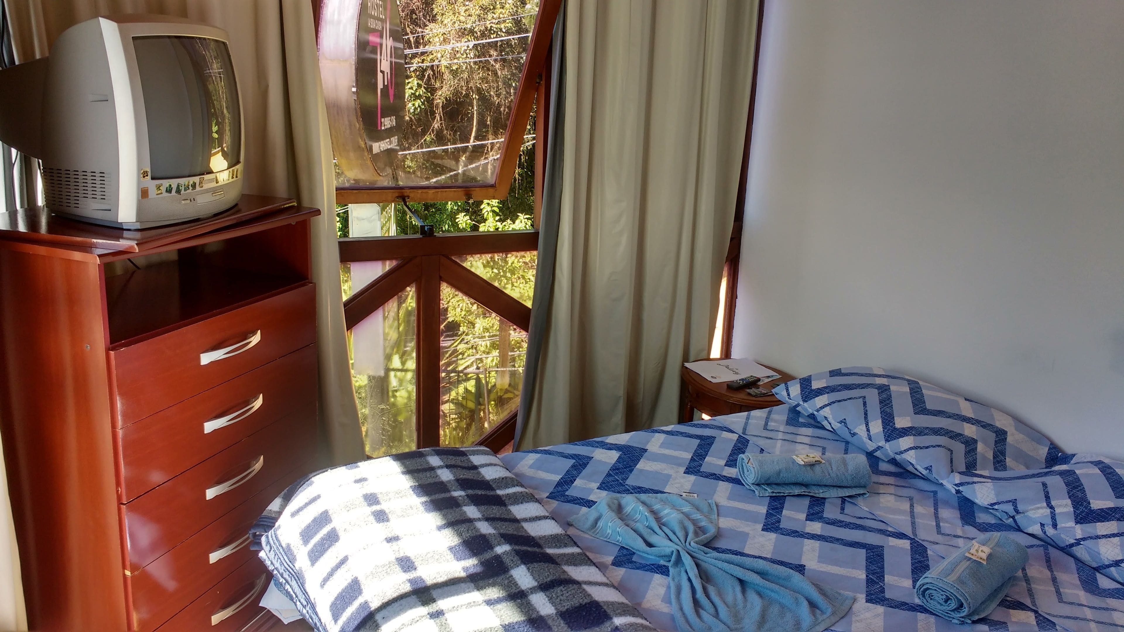 A bedroom with a bed, a wooden dresser, a television on top, and a window with curtains.