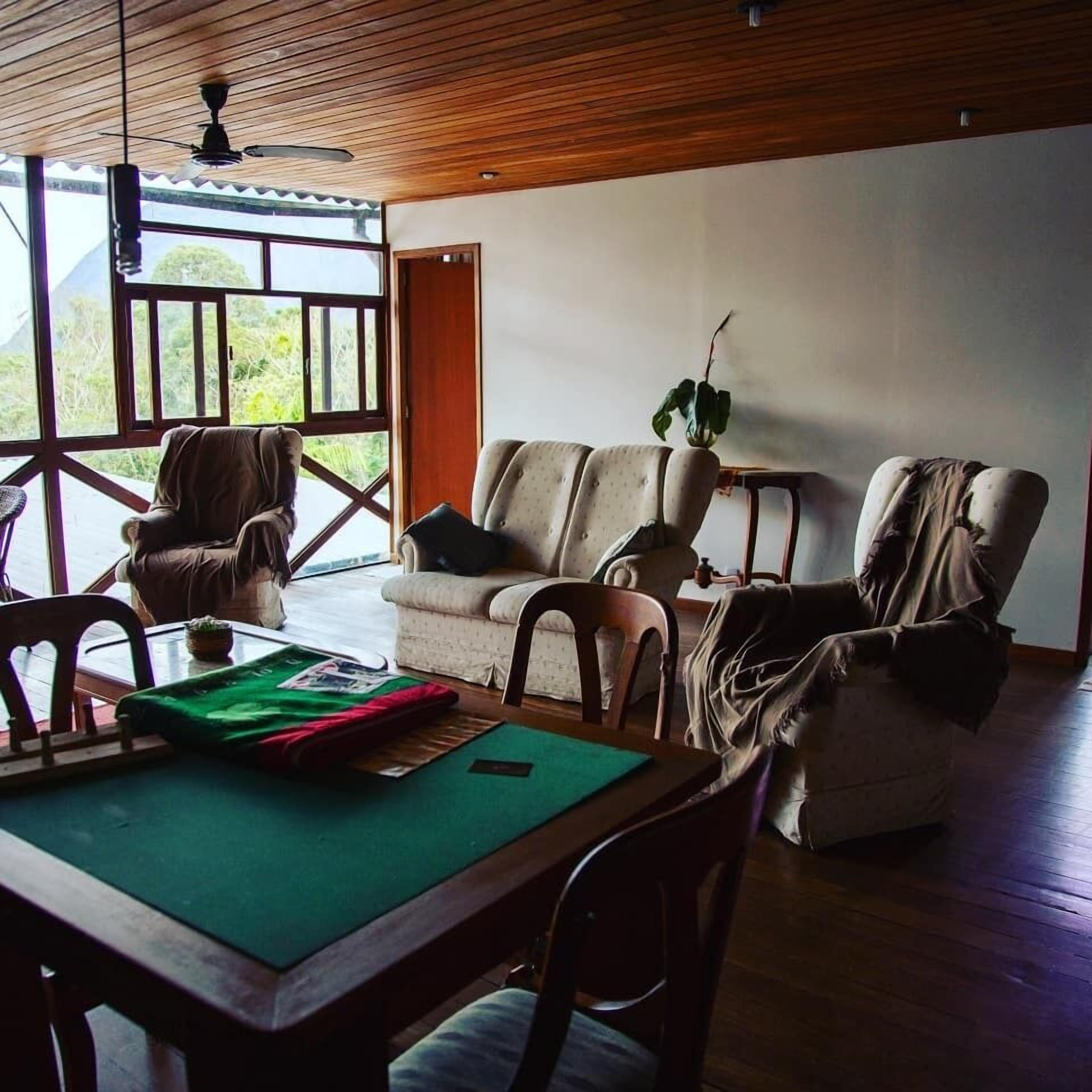 A room with a wooden ceiling, a sofa, a chair, a dining table with a green placemat, and a view through large windows.