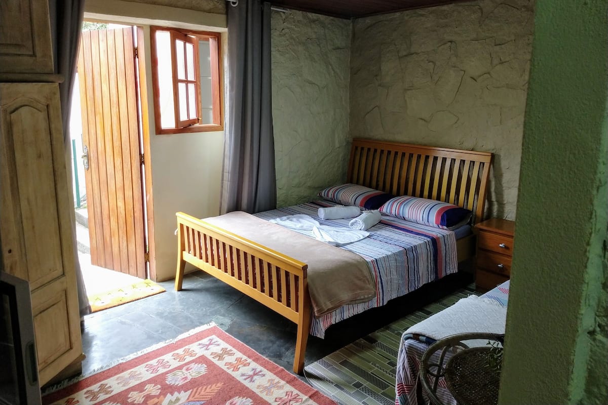 Un dormitorio con cama de madera, una mesita de noche, una pequeña mesa, una silla, una ventana con cortinas y una puerta.