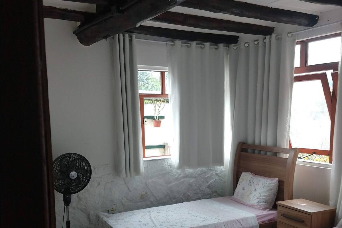 Una habitación pequeña y acogedora con dos camas individuales, una mesita de noche de madera, un buró, un ventilador y una ventana con cortinas.