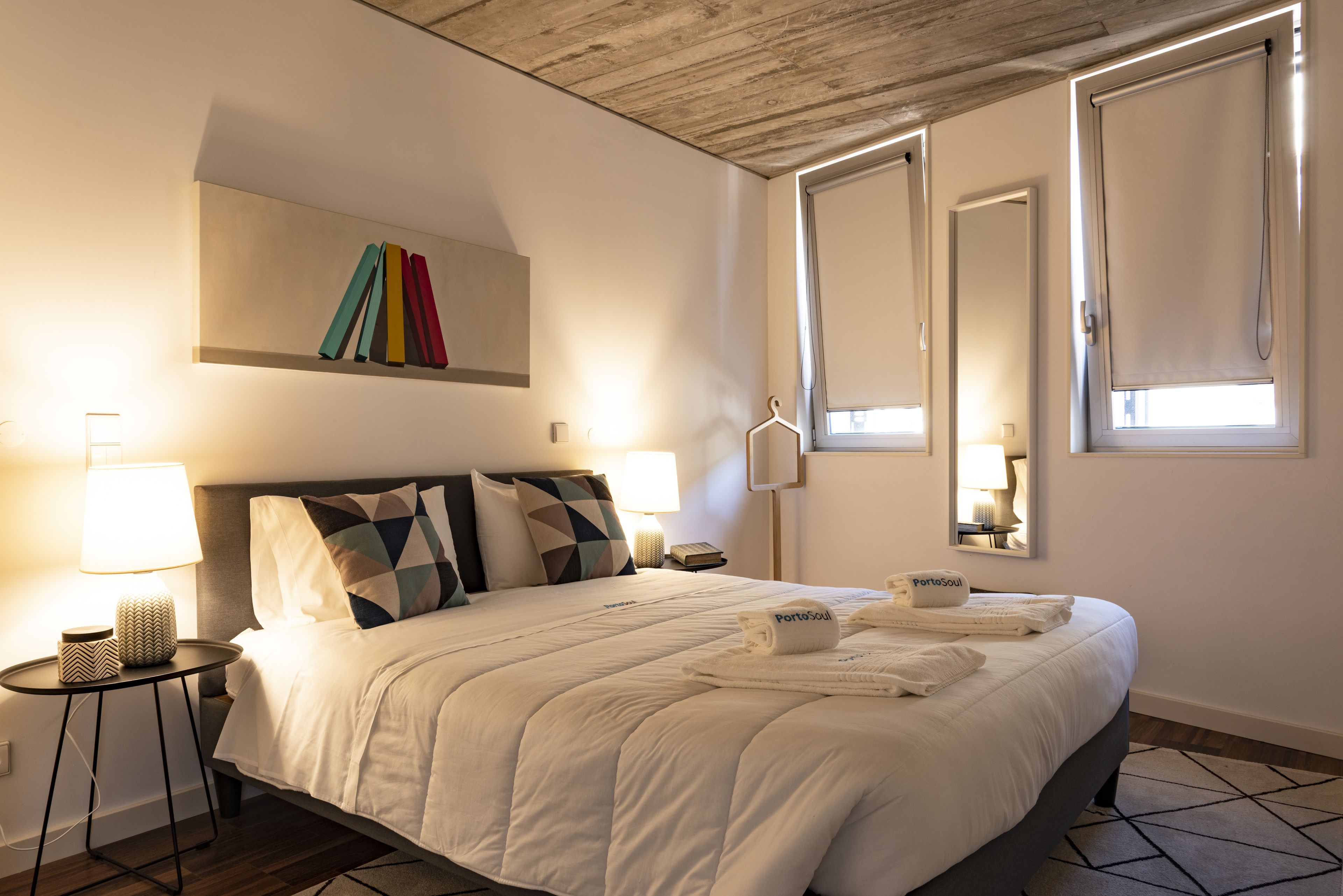 Una cama bien hecha con sábanas blancas y una toalla PortoSoul, una mesita de noche con una lámpara, y una obra de arte colgada en la pared.