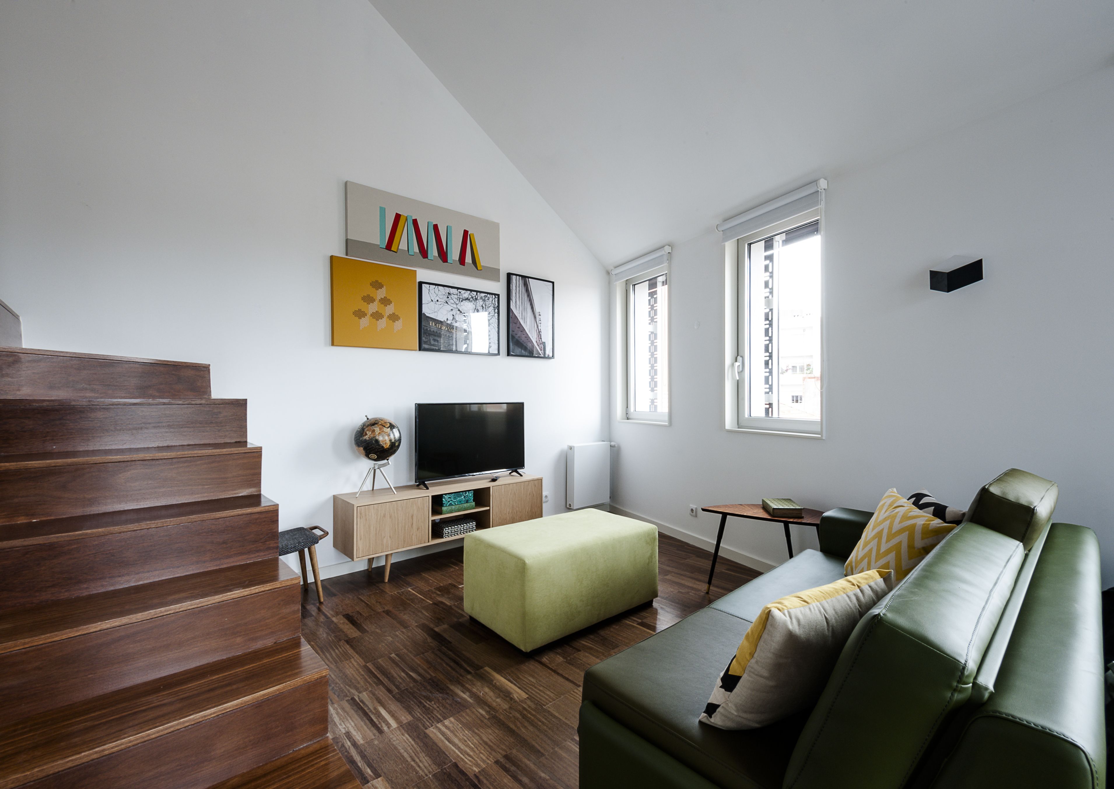 A modern living room with a staircase, a green sofa, a TV on a wooden stand, and artwork on the walls.