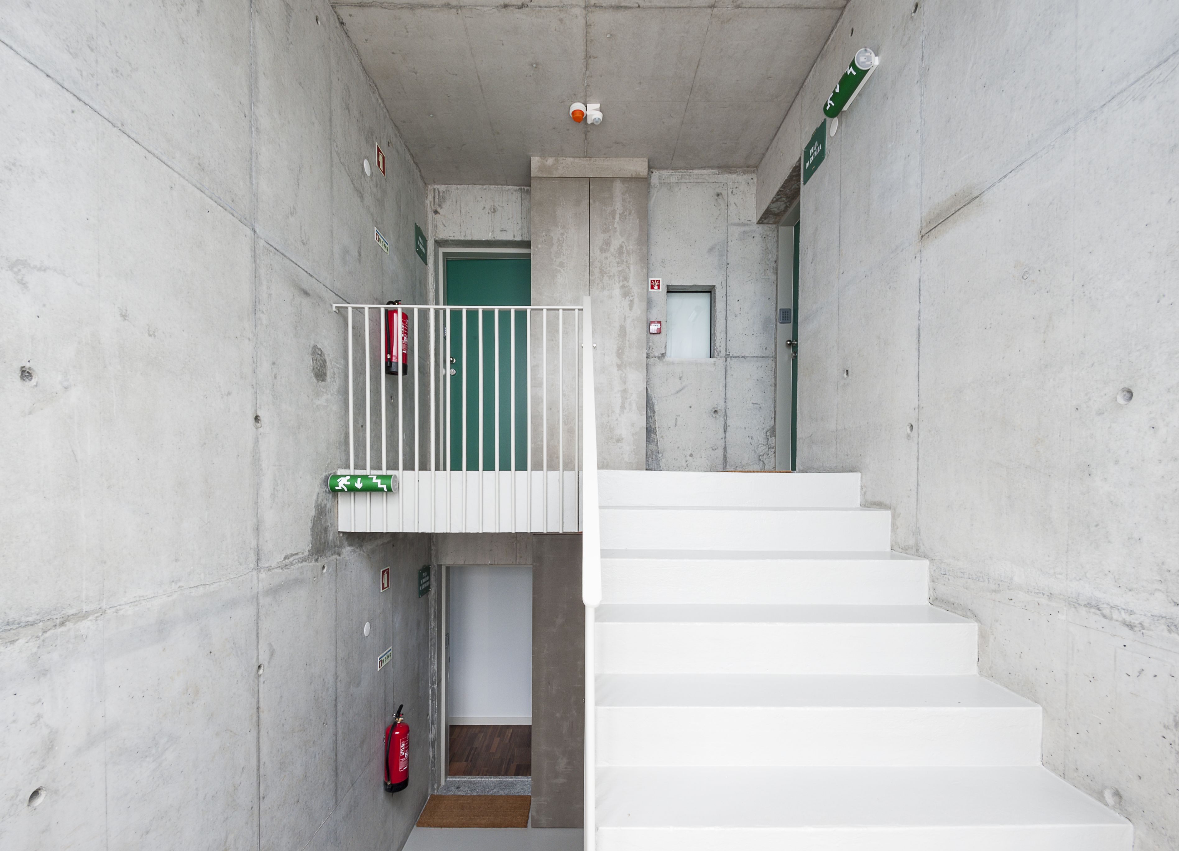 Un escalier en béton, avec une porte verte en haut et un extincteur fixé au mur.