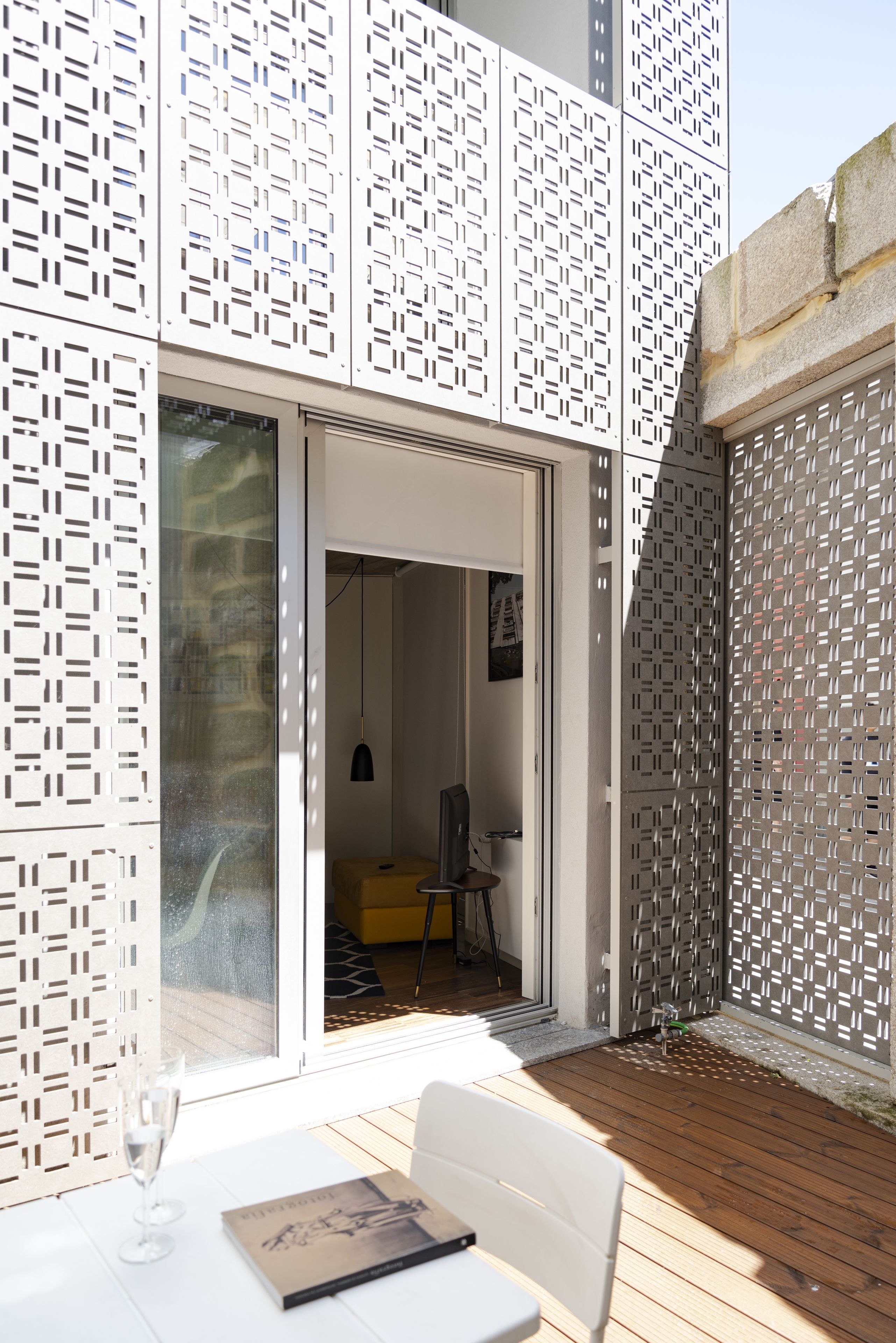 Patio esterno con tavolo, un bicchiere d'acqua e un libro. Una porta scorrevole in vetro dà accesso a un ambiente interno con un letto e una lampada.