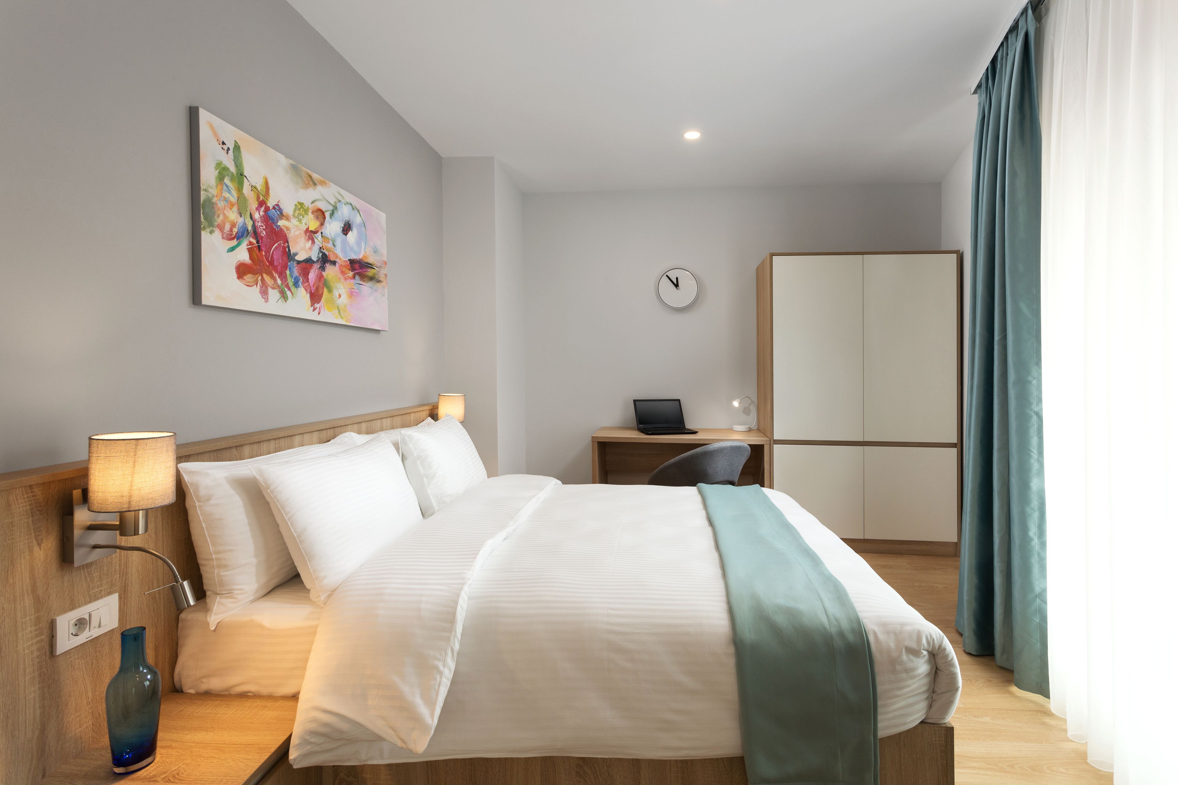 Una camera d'albergo con un letto, lampade ai comodini, una scrivania con un portatile, una sveglia e un armadio.