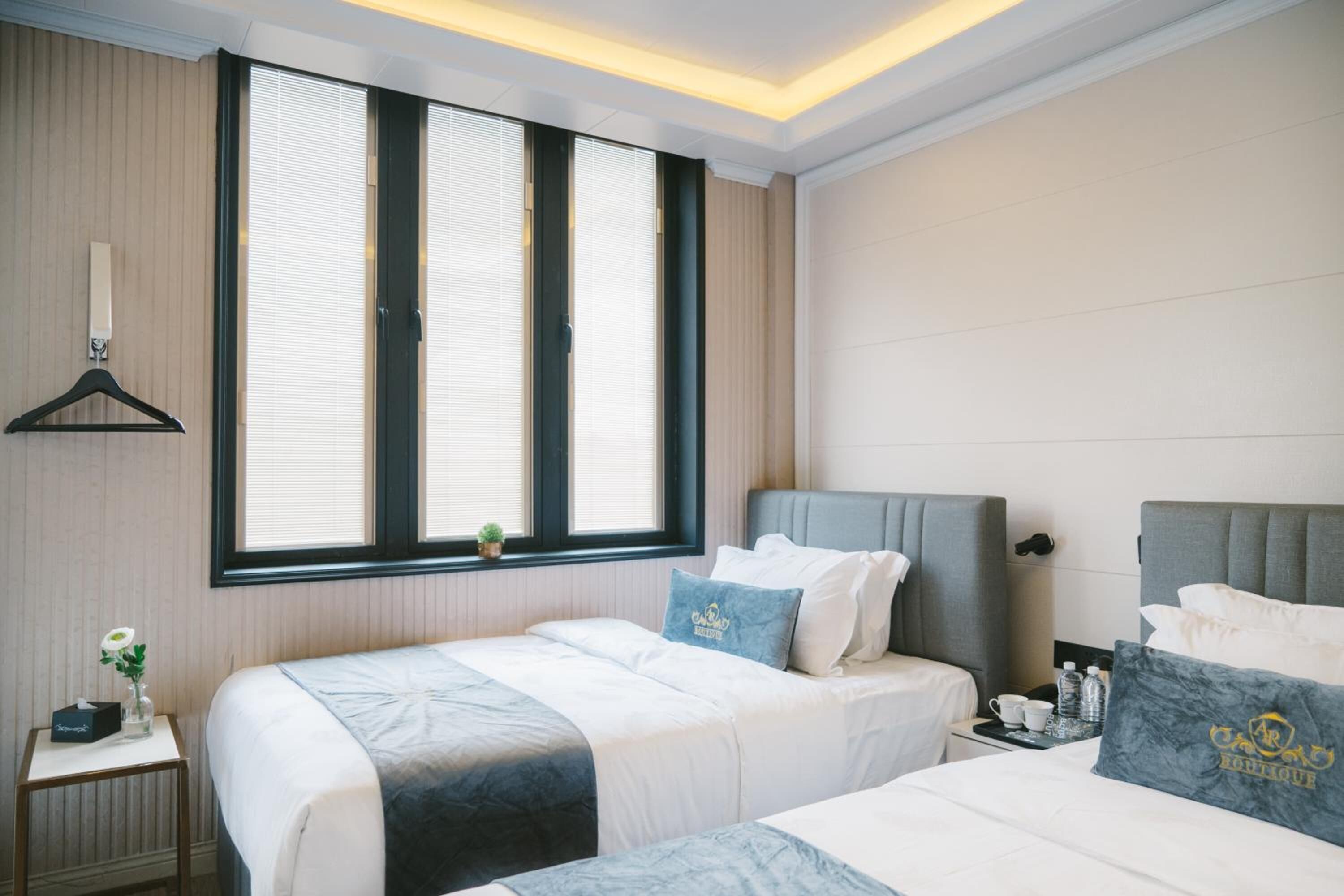 Habitación de hotel con dos camas, una mesita con una planta, una ventana con persianas y una mesita de noche con una taza y botellas de agua.